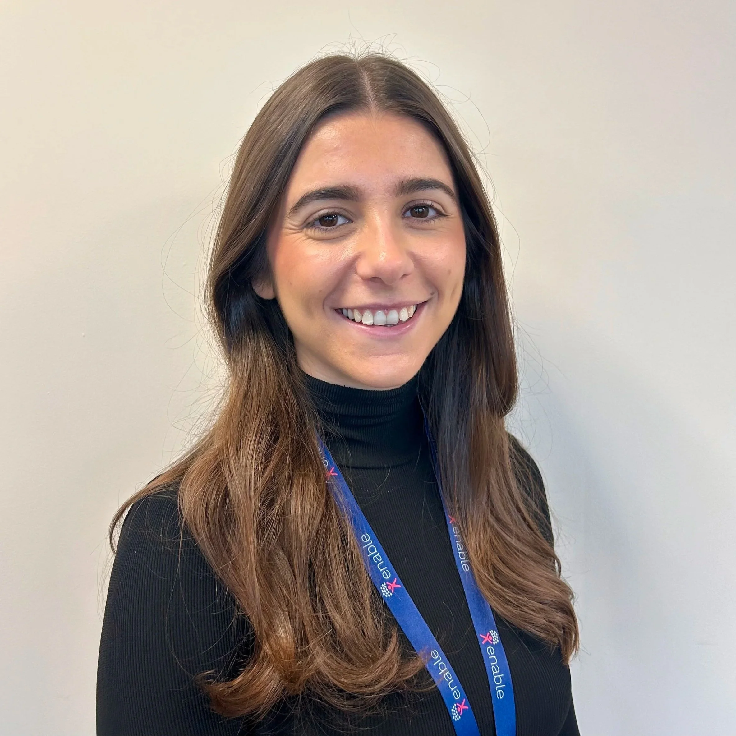 Woman smiling with long dark brown hair, wearing a blue 'Enable' lanyard, against a blank wall.