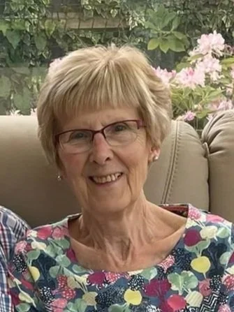 An older lady with short blonde hair and glasses smiling wearing a floral top, sat down on the sofa.