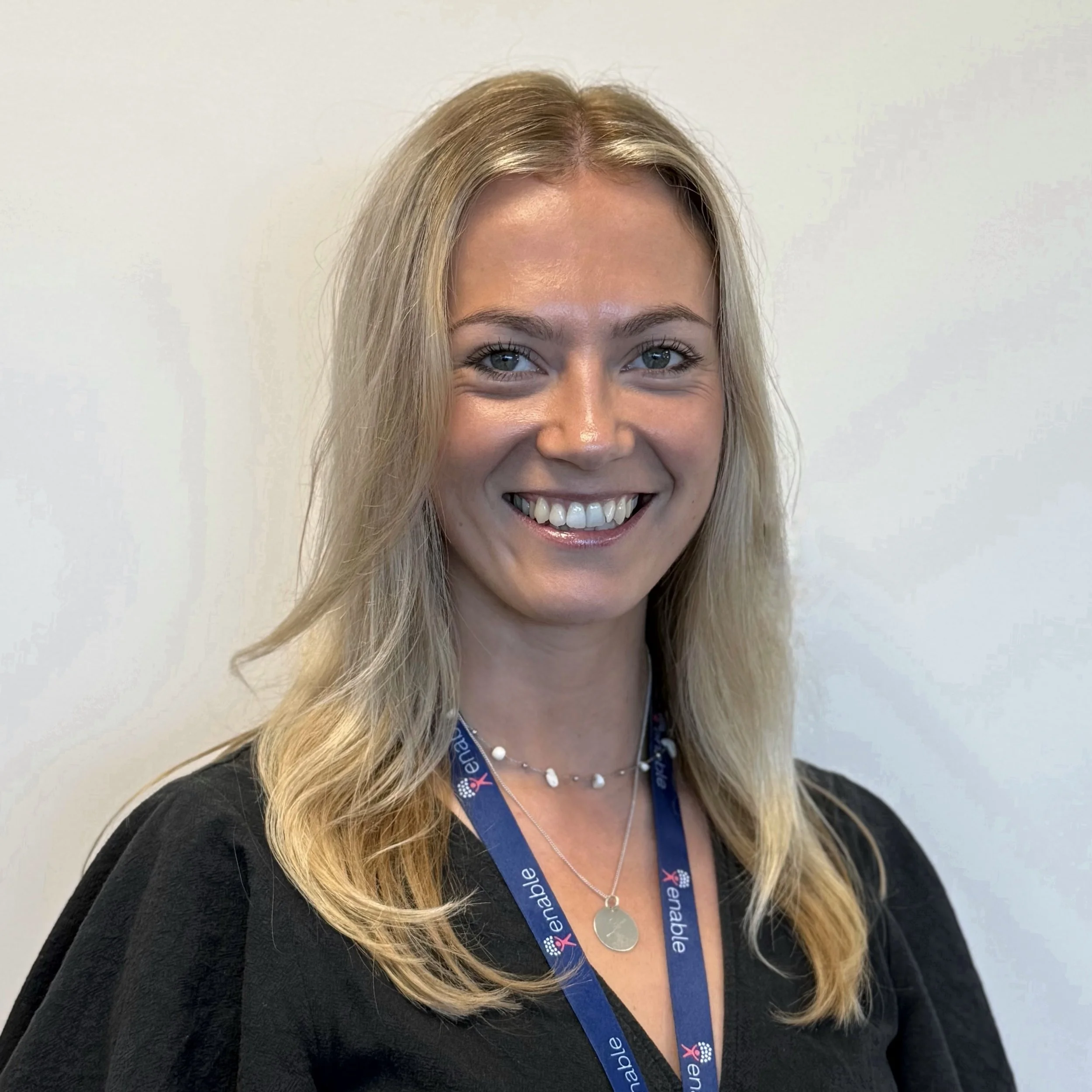 Shoulder length blonde hair, wearing a black top and a blue lanyard, smiling in front of a plain wall.