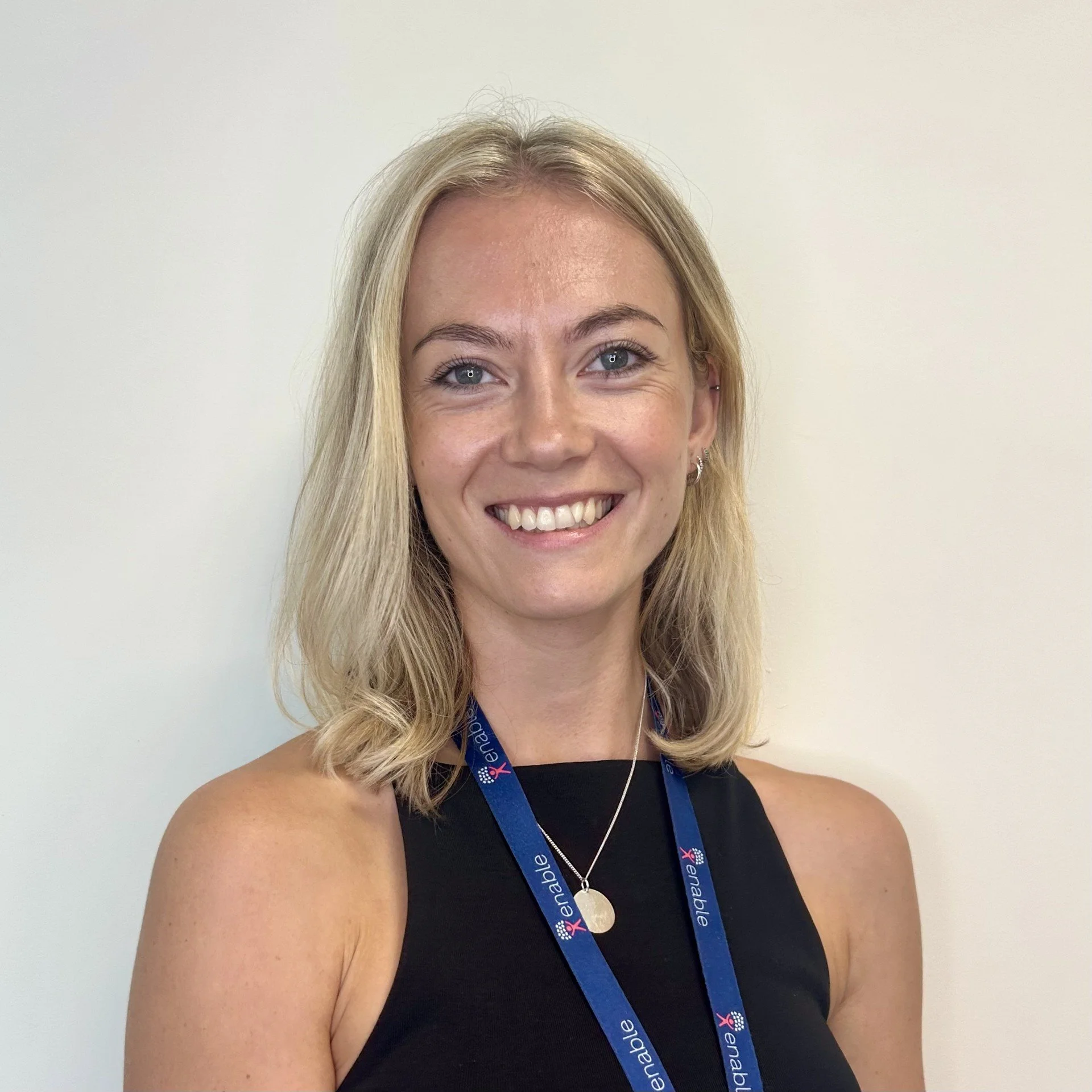 Shoulder length blonde hair, wearing a black top and a blue lanyard, smiling in front of a plain wall.