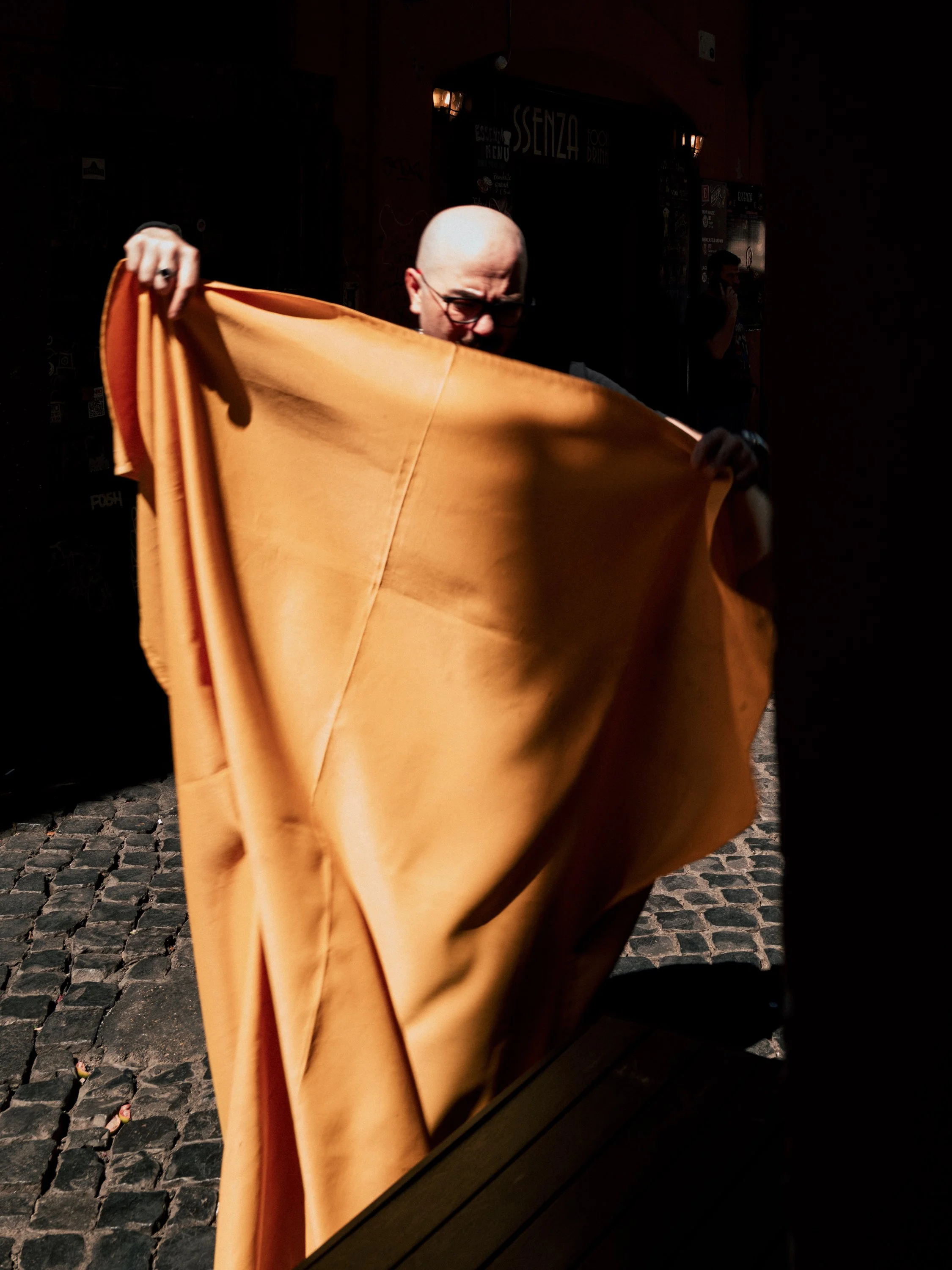 A person with glasses and a bald head holding a large orange fabric in front of their face. The person is outdoors on a cobblestone street with signs in the background including one that says 'Senza'.