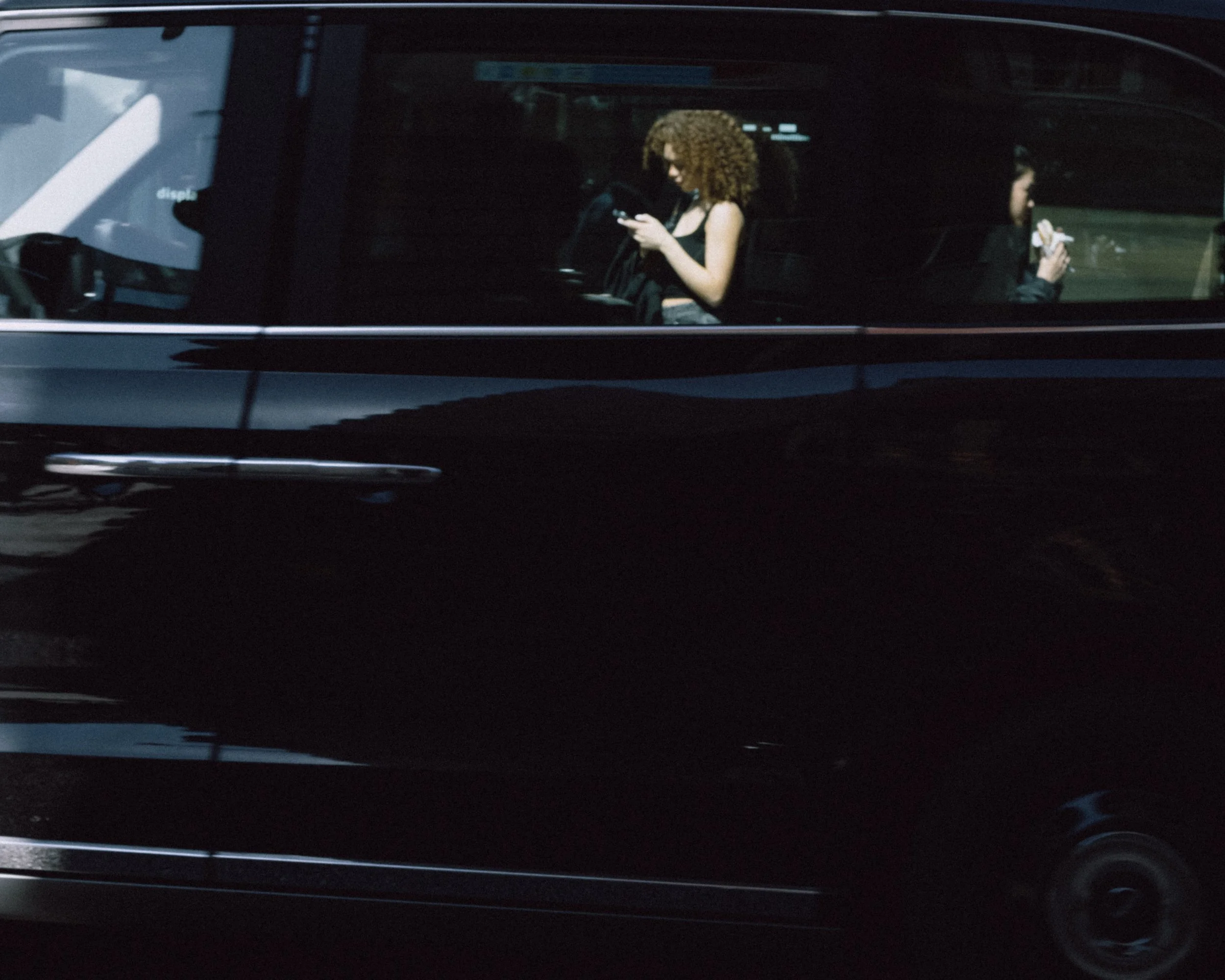 Two women sitting inside a black vehicle, each using a cellphone.