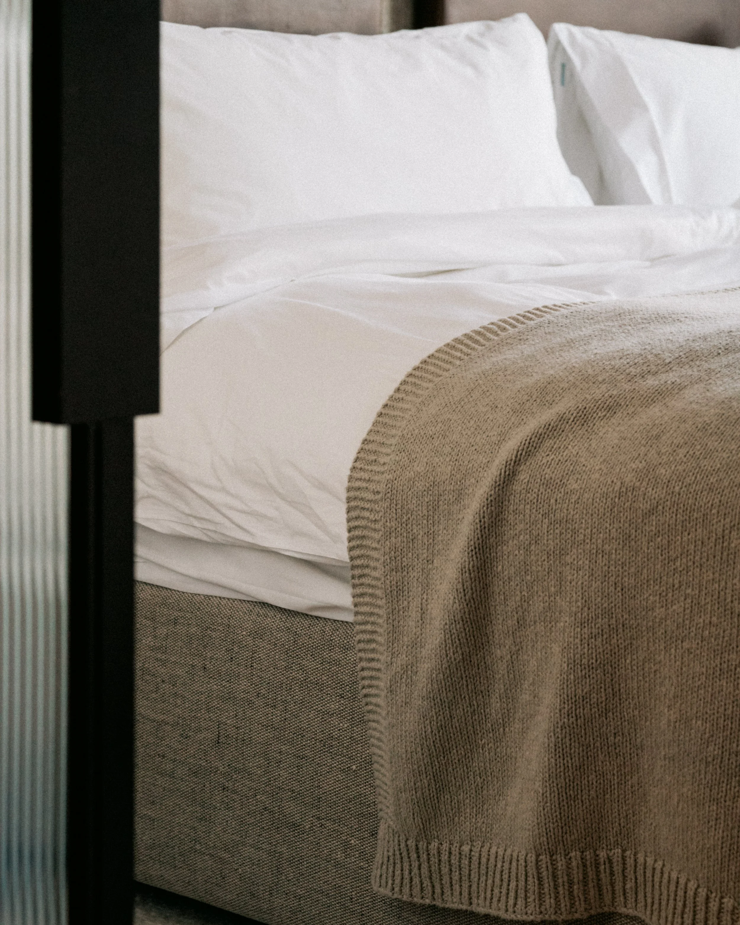 A neatly made bed with white sheets, two white pillows, and a beige knitted blanket at the foot of the bed in a bedroom.