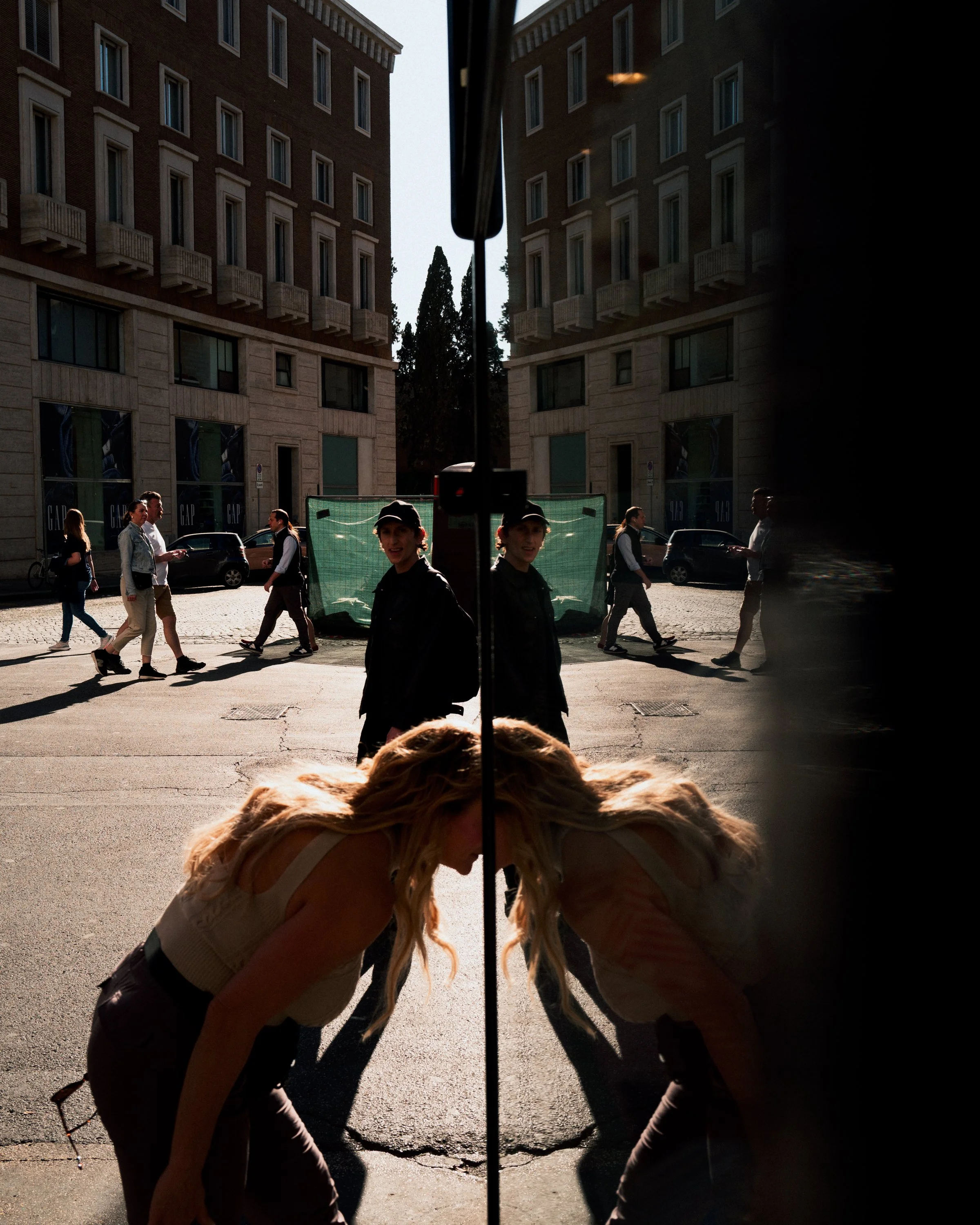 A woman with blonde hair bending down in front of a large glass door with reflection. The reflection shows people walking on the street outside, two women in black caps in the foreground, and multi-story buildings.