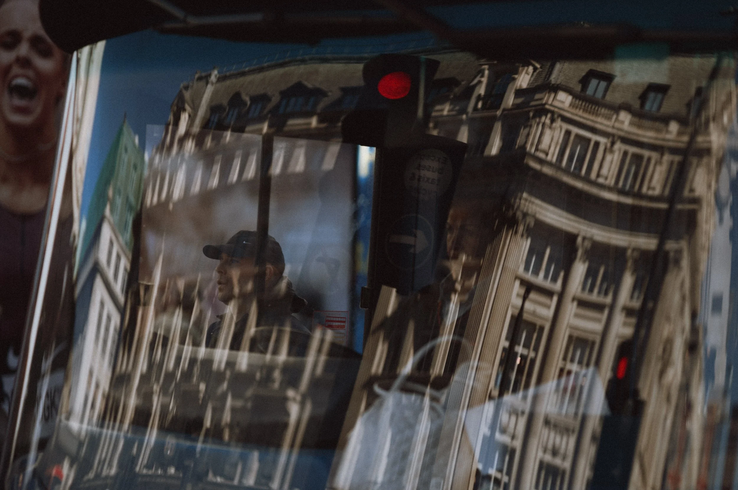 Reflection of a building with classical architecture in a bus window, with a man wearing a cap and sunglasses inside the bus.