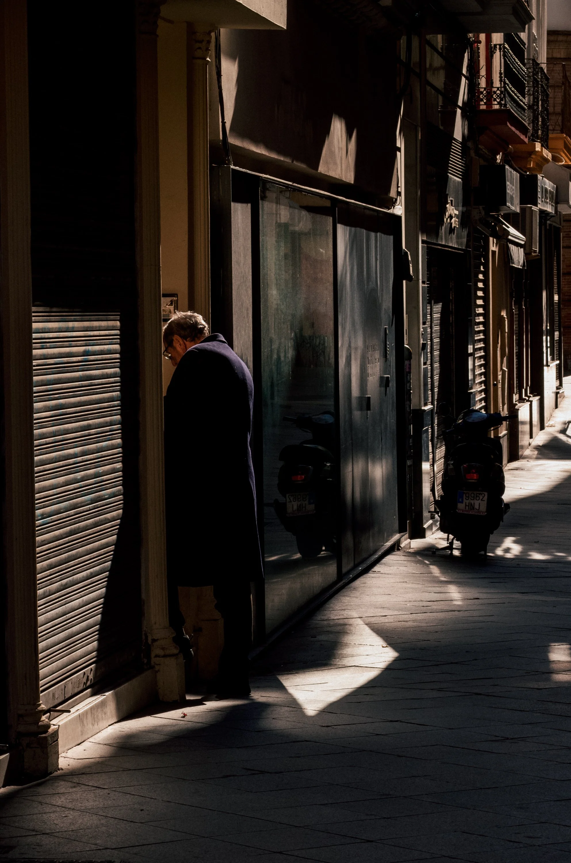A man with glasses and a dark coat leaning against a closed storefront, partially in shadow, on a city sidewalk with parked scooters visible.