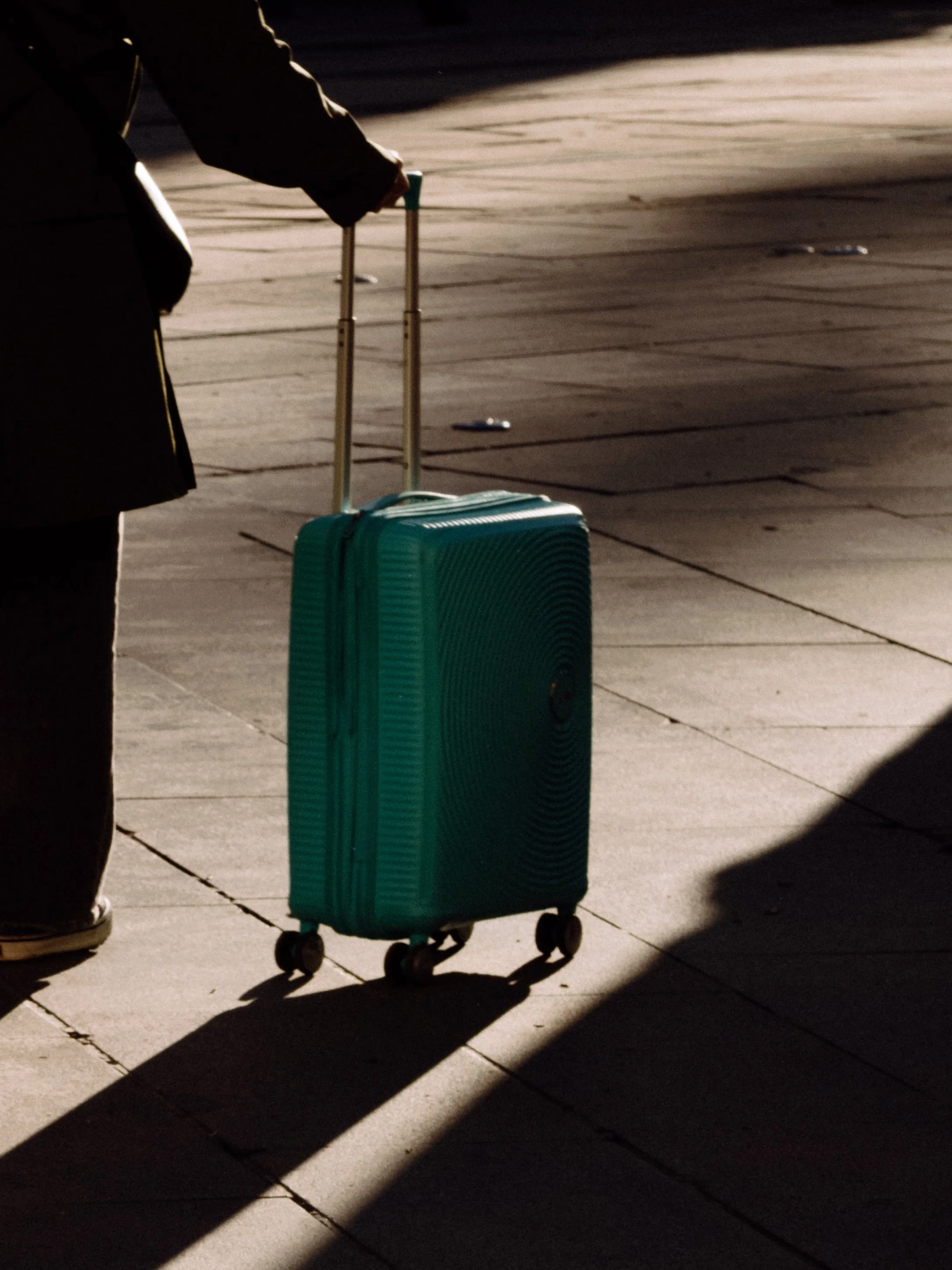 Person pulling teal suitcase on airport or train station platform with shadows and sunlight.