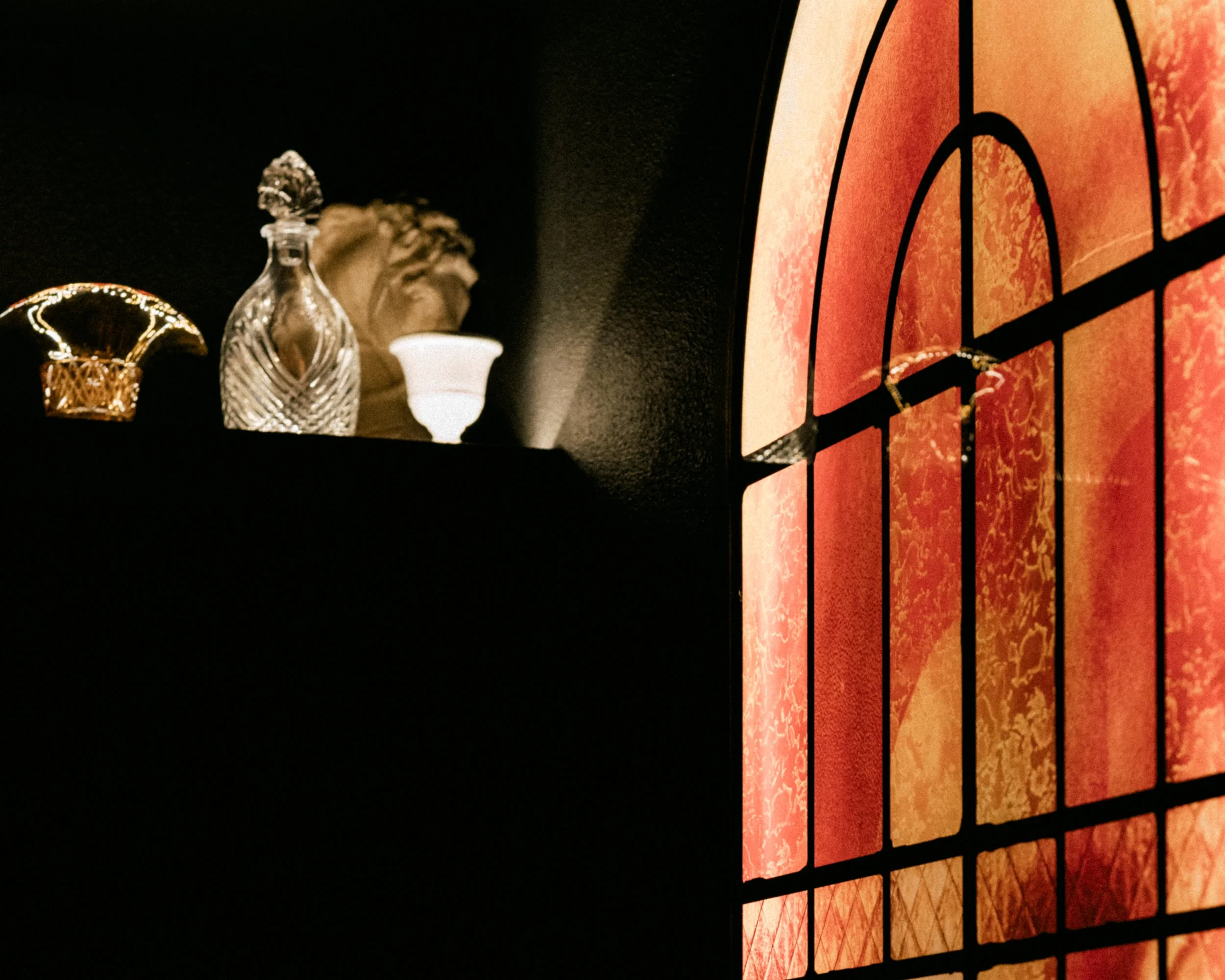 Close-up of a dartboard with orange and red sections, black dividing lines, and a black background. Various glass decanters and a lamp are in the foreground, slightly out of focus.