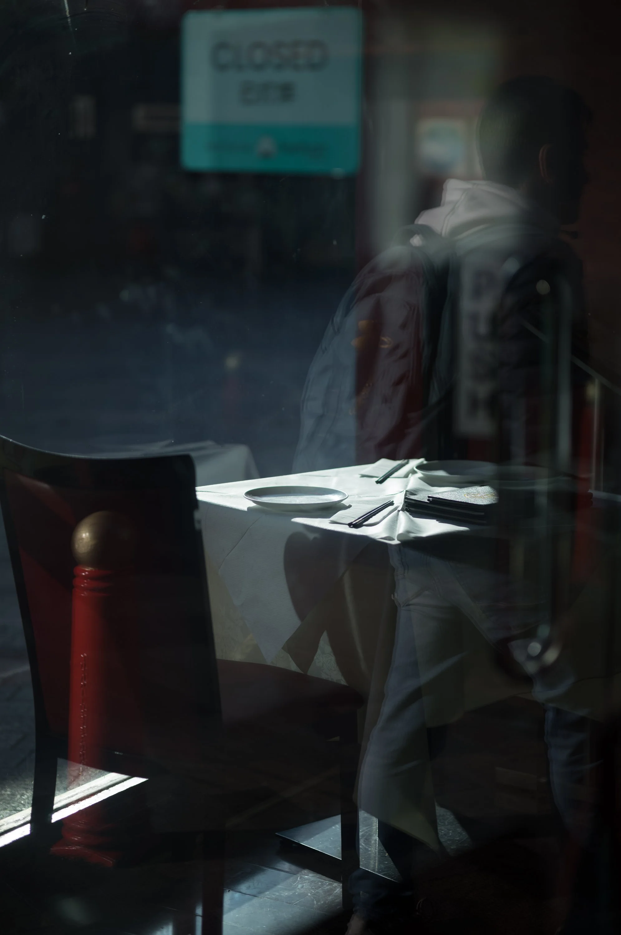 Reflection of a person sitting at a table with a white tablecloth in a restaurant seen through a window, with a sign in the background that reads 'Closed'.