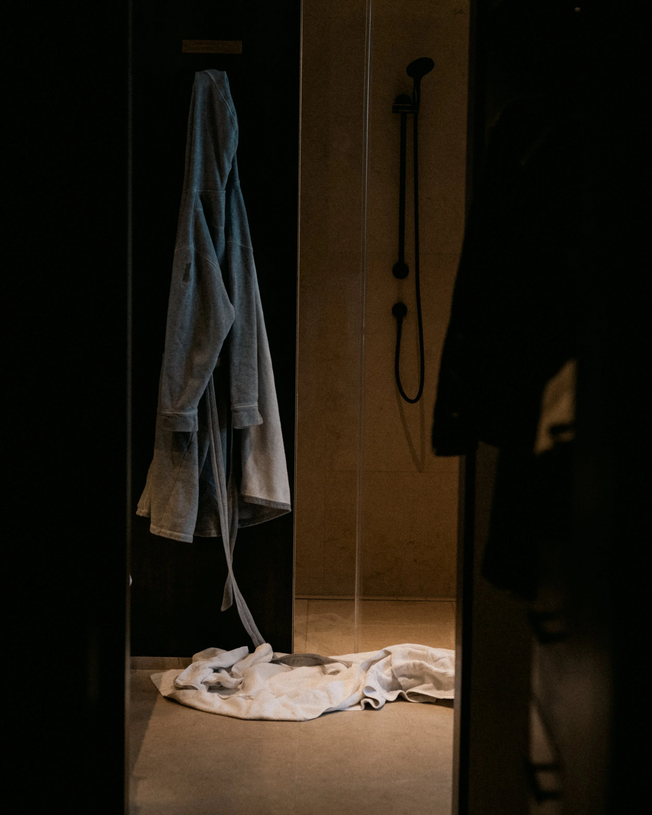 A dark reflection of a doorway with a gray bathrobe hanging inside, a peephole on the door, a wall-mounted shower head, a towel on the floor, and a person’s silhouette outside.