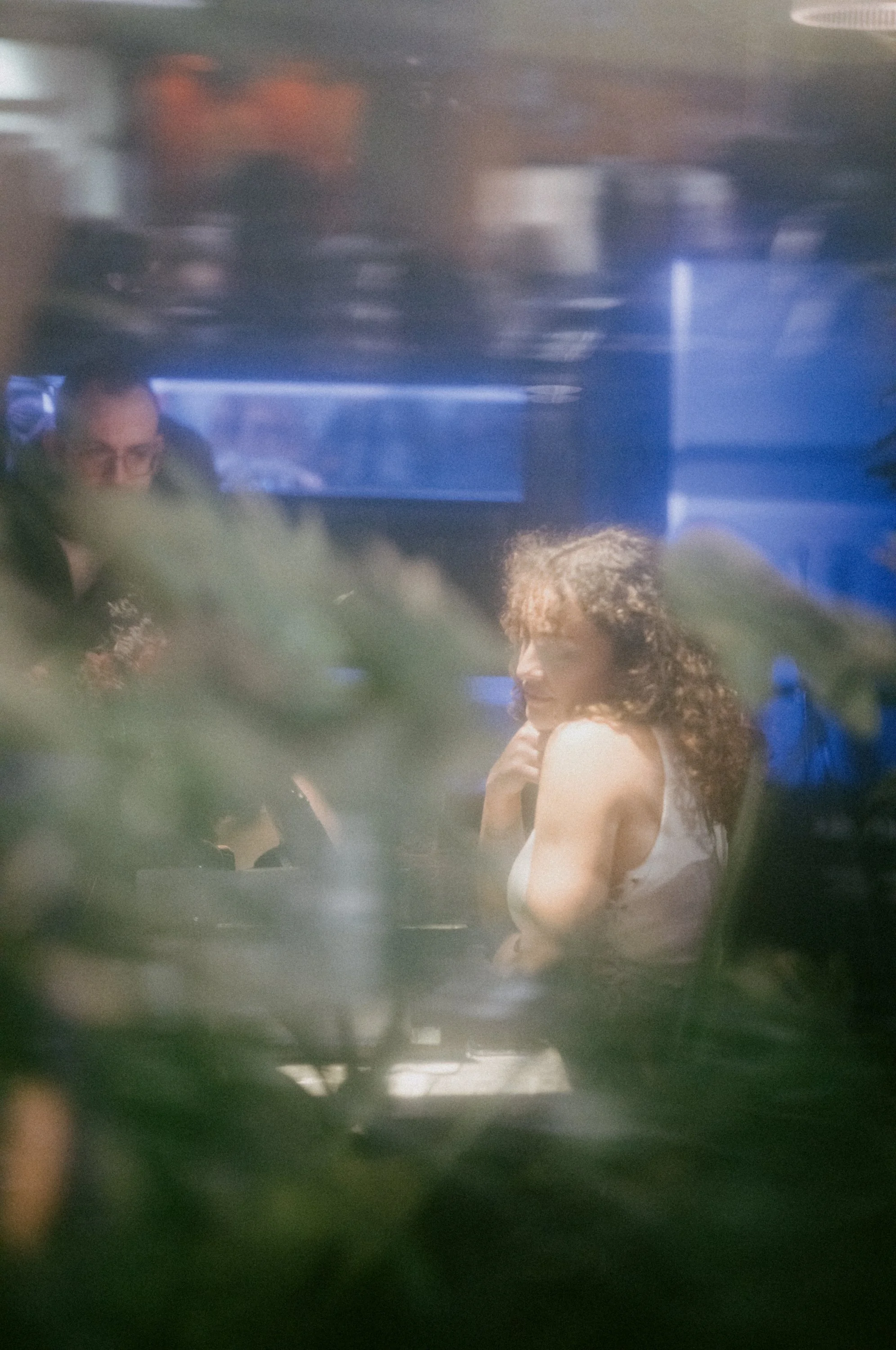 A woman with curly hair and a white sleeveless top sitting at a table, with a man nearby, in a shaded or blurry setting with greenery in the foreground.