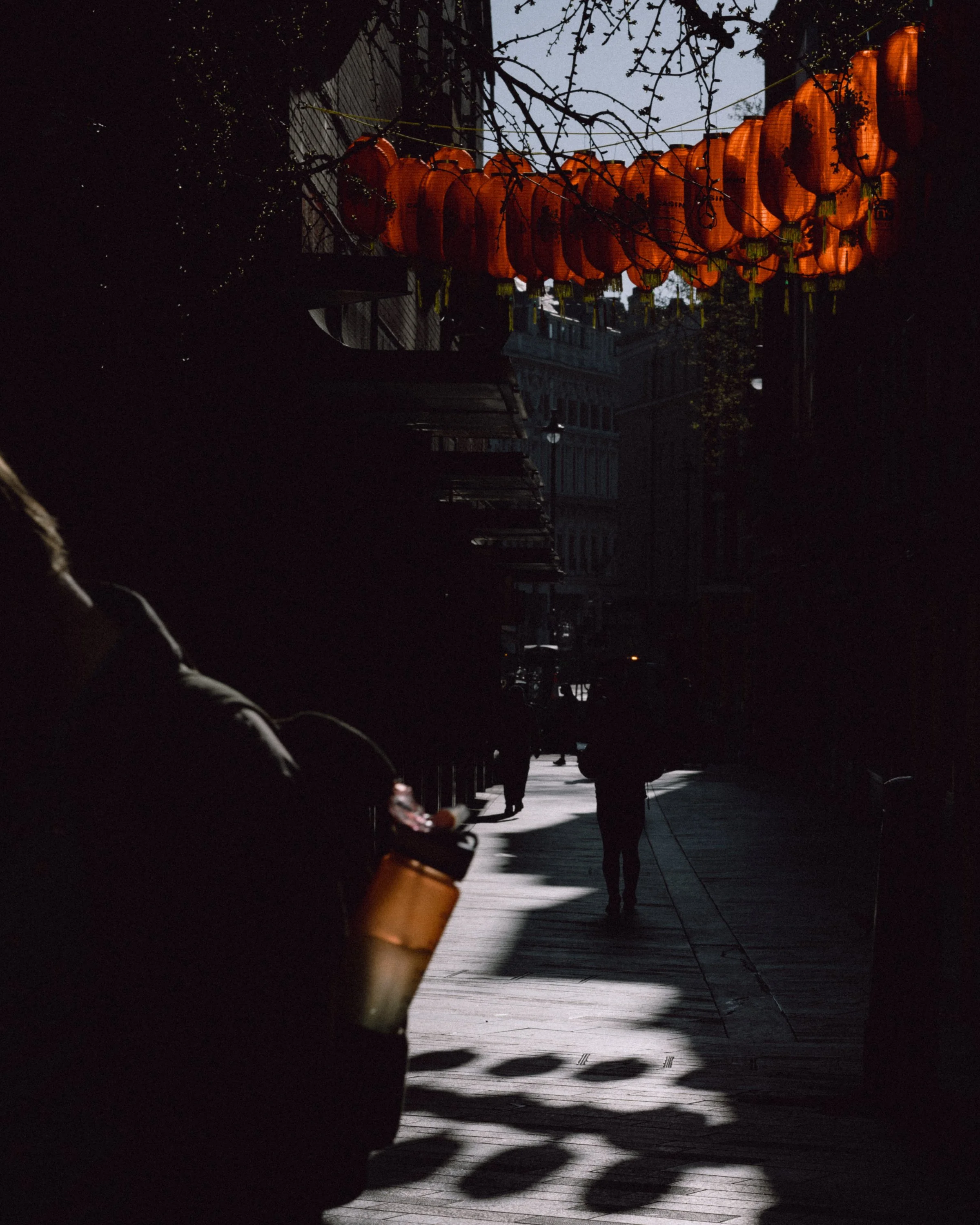 Orange lanterns hanging above a dark street with silhouetted people walking and shadows on the sidewalk.