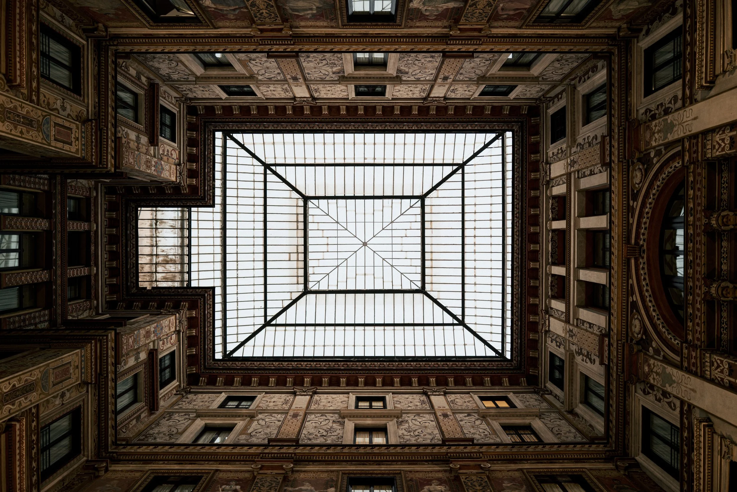 View looking up at an ornate glass ceiling inside a historic building surrounded by decorated walls with multiple windows.