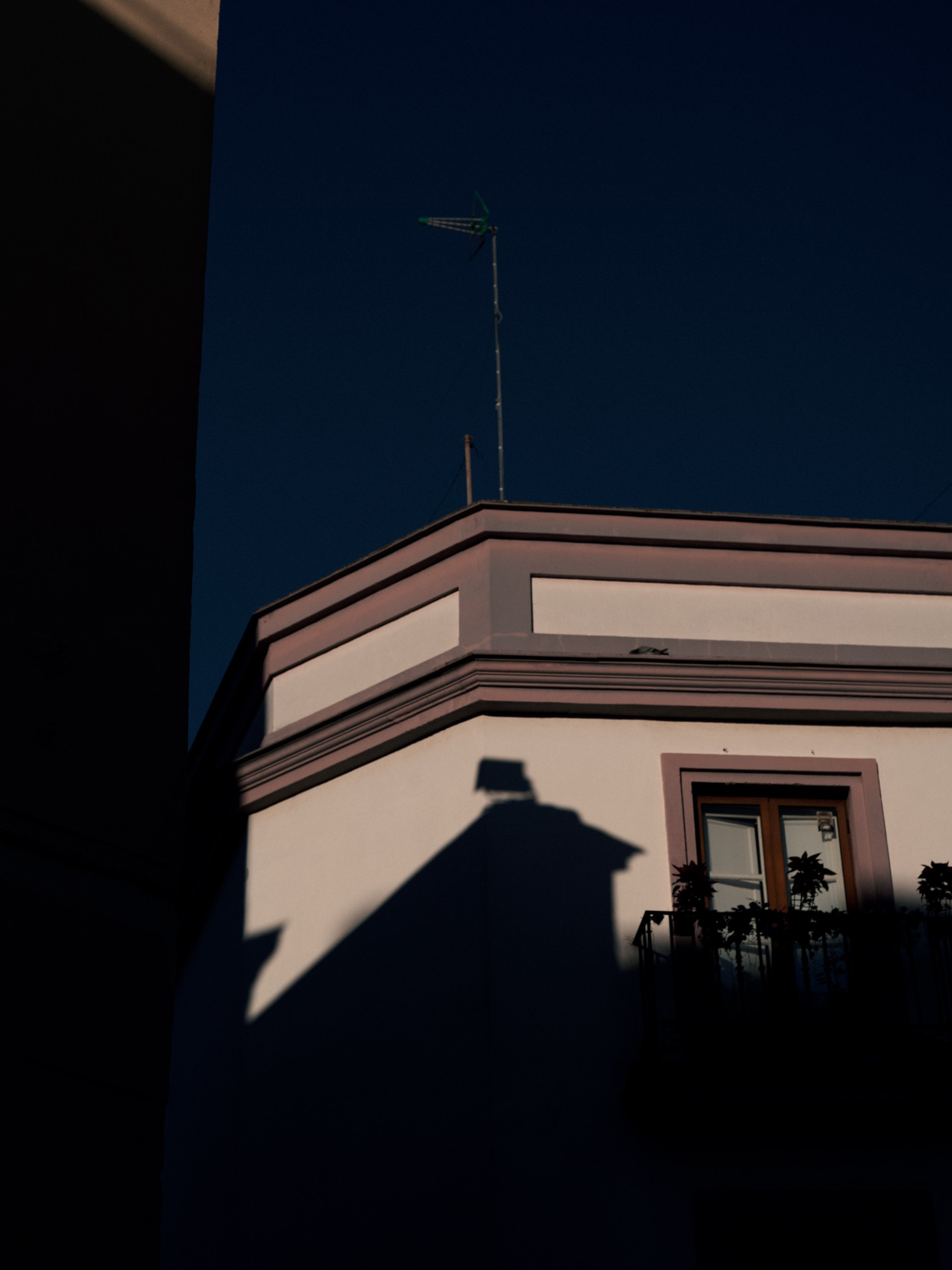 Shadow of a building's roof and window cast on an exterior wall with a balcony and potted plants, against a dark evening sky with TV antenna on the roof.