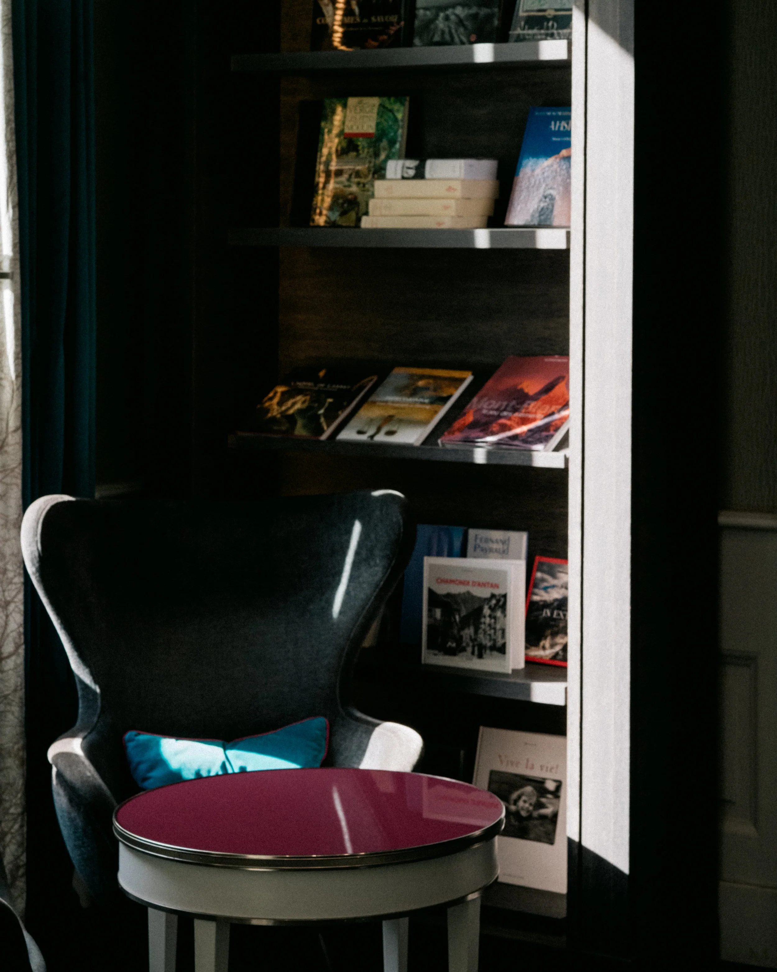 A bookshelving unit filled with books, magazines, and papers, with a dark wood finish, located in a room with a curtain on the left and a black upholstered chair nearby. In front, there is a round table with a pink and white top, next to a black chai