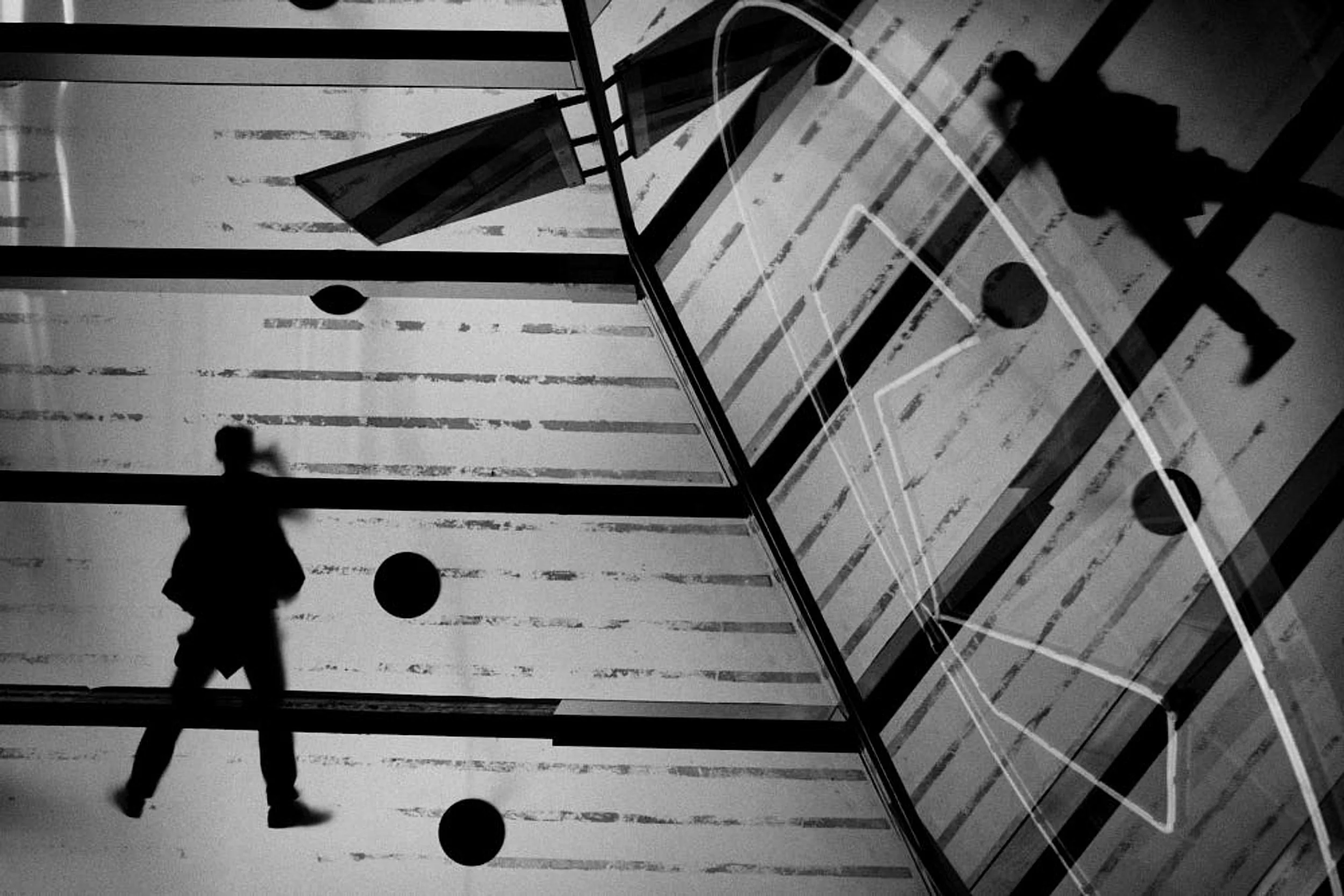 Silhouettes of two people walking on a glass floor with reflections and round black dots, overlaid with an outline of a soccer field on the glass surface.