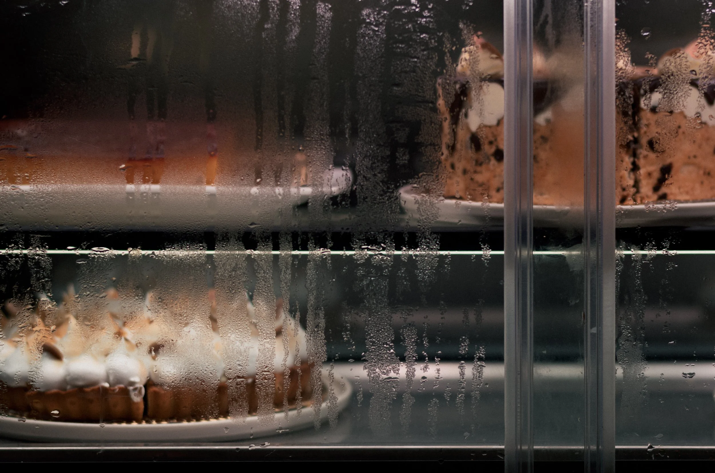 Desserts on display behind a foggy glass shelf, including a cupcake with whipped topping and chocolate drizzle, and a sliced chocolate cake.