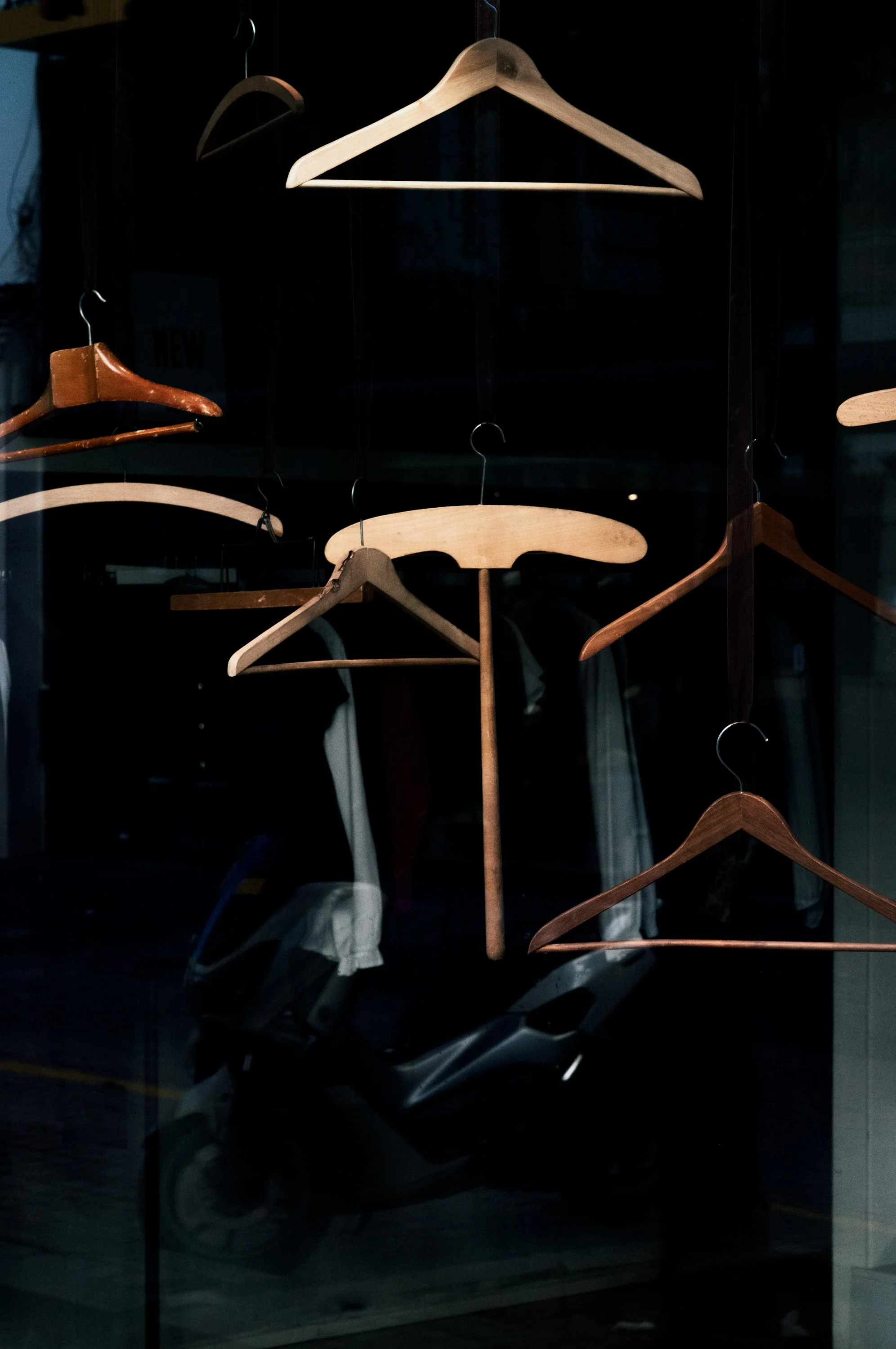 Multiple wooden hangers hanging in a dark retail window display with a reflection of a motorcycle outside.