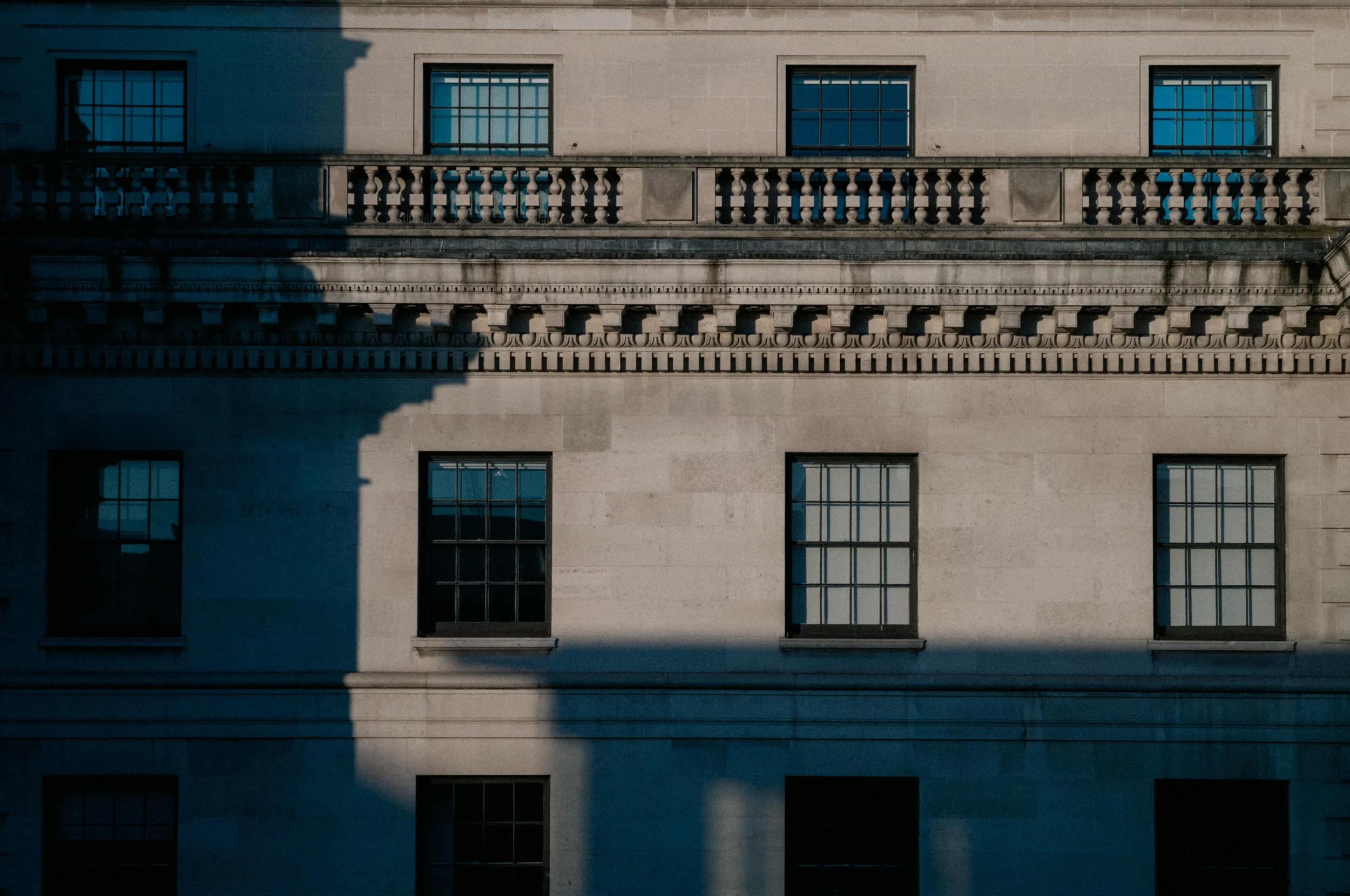The image shows the front of a building with multiple windows and decorative stonework. Shadows of a tree are cast across the facade.