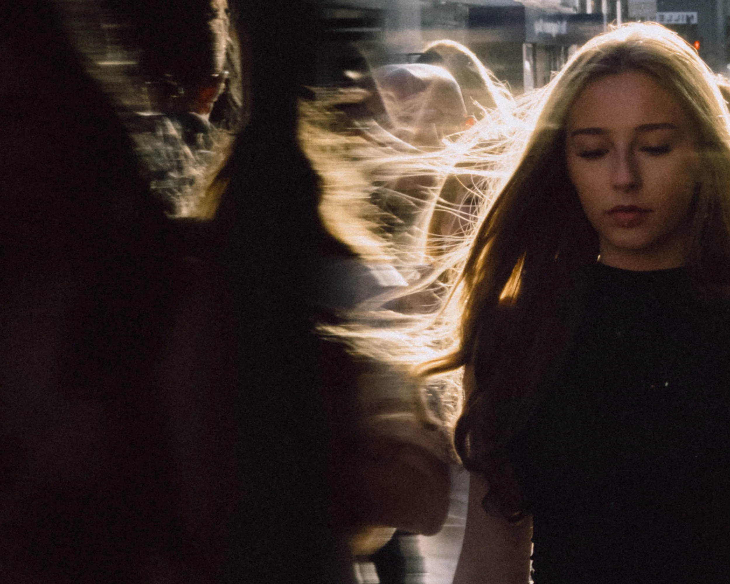 A woman with long blonde hair stands outdoors at night, with sunlight illuminating her hair. Other people are visible in the blurred background.