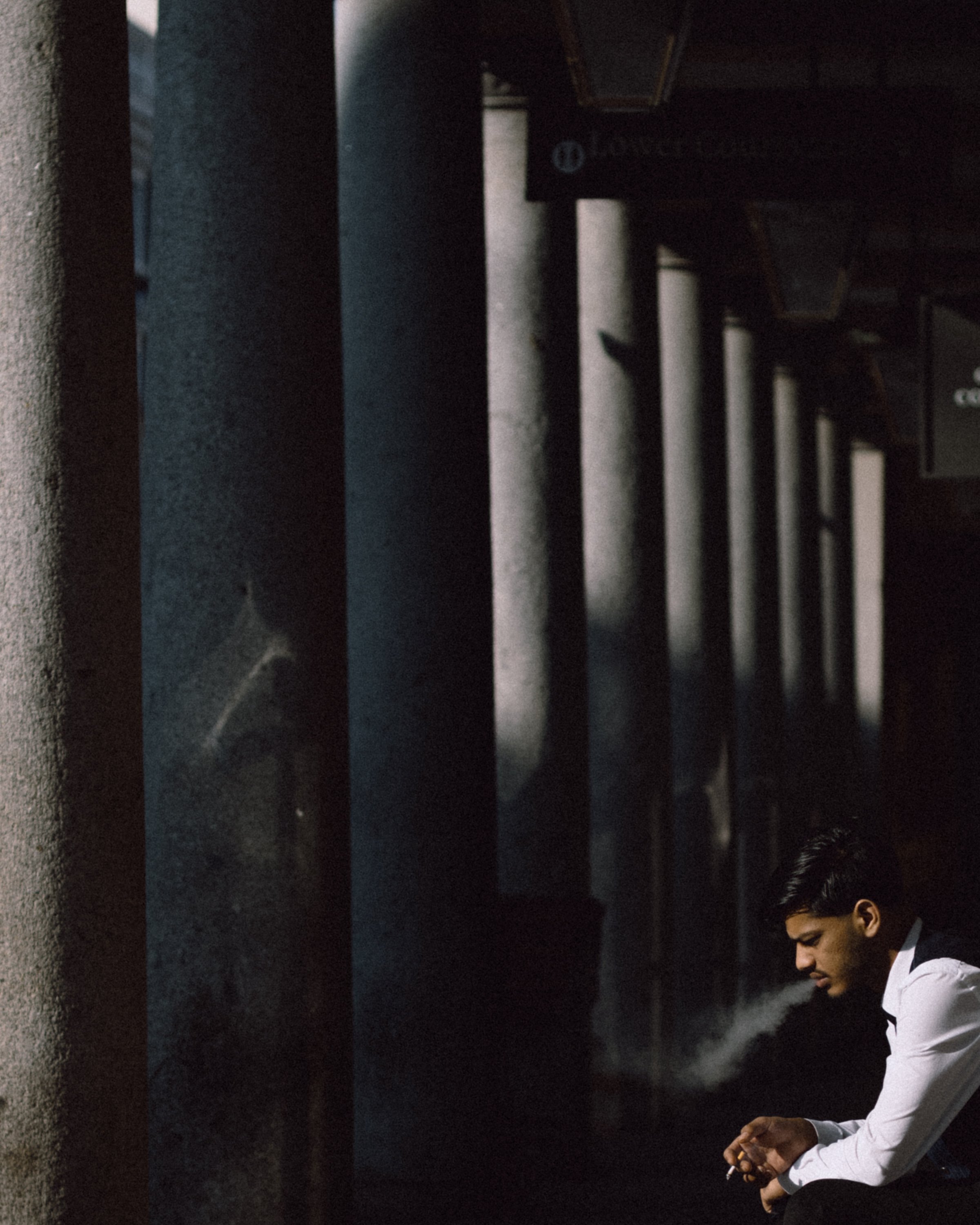 A young man sitting in an outdoor area, appears to be smoking and looking at his phone, with vertical concrete pillars in the background.