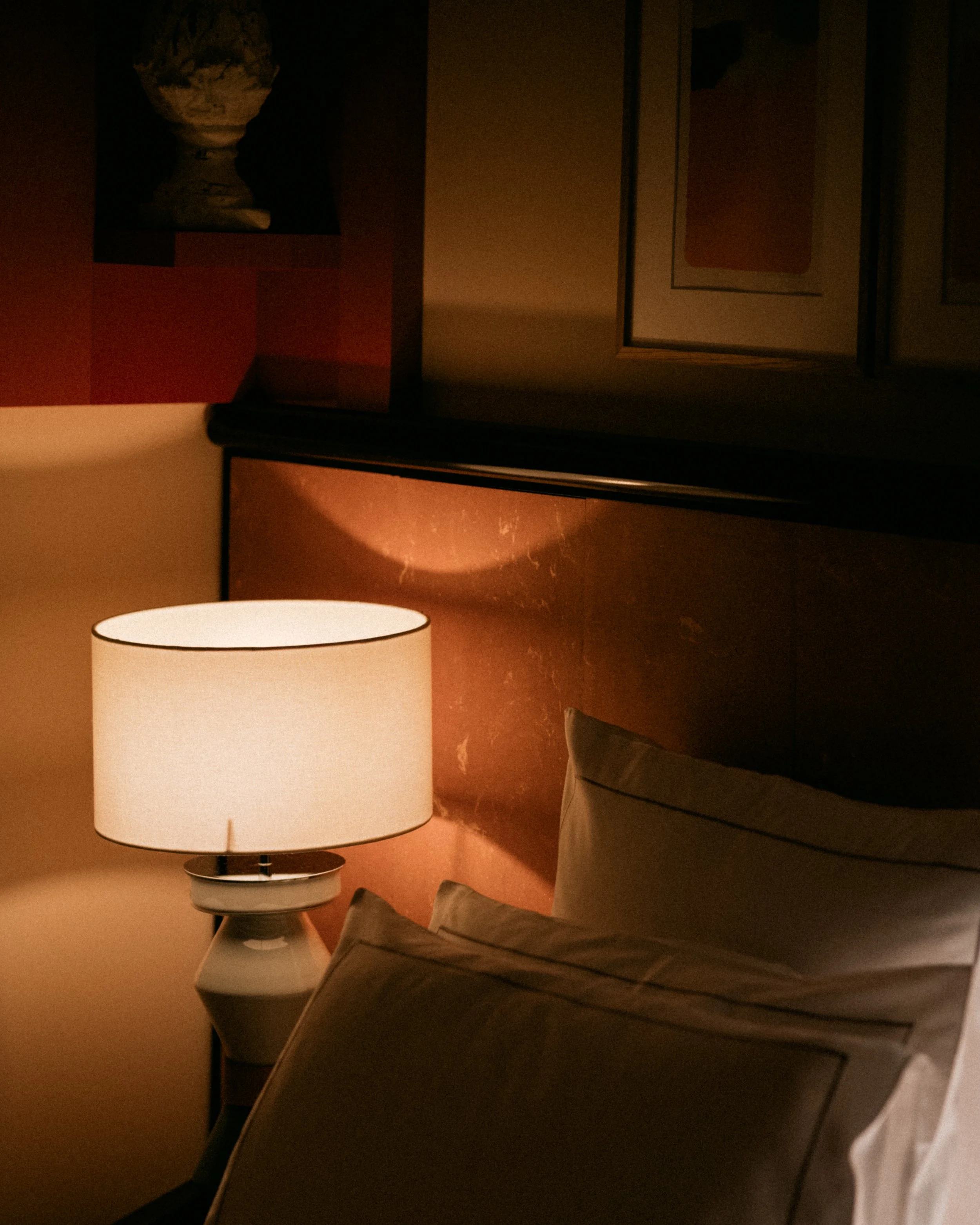 A person sitting on a brown couch with their legs crossed, next to a white table lamp in a dimly lit room.