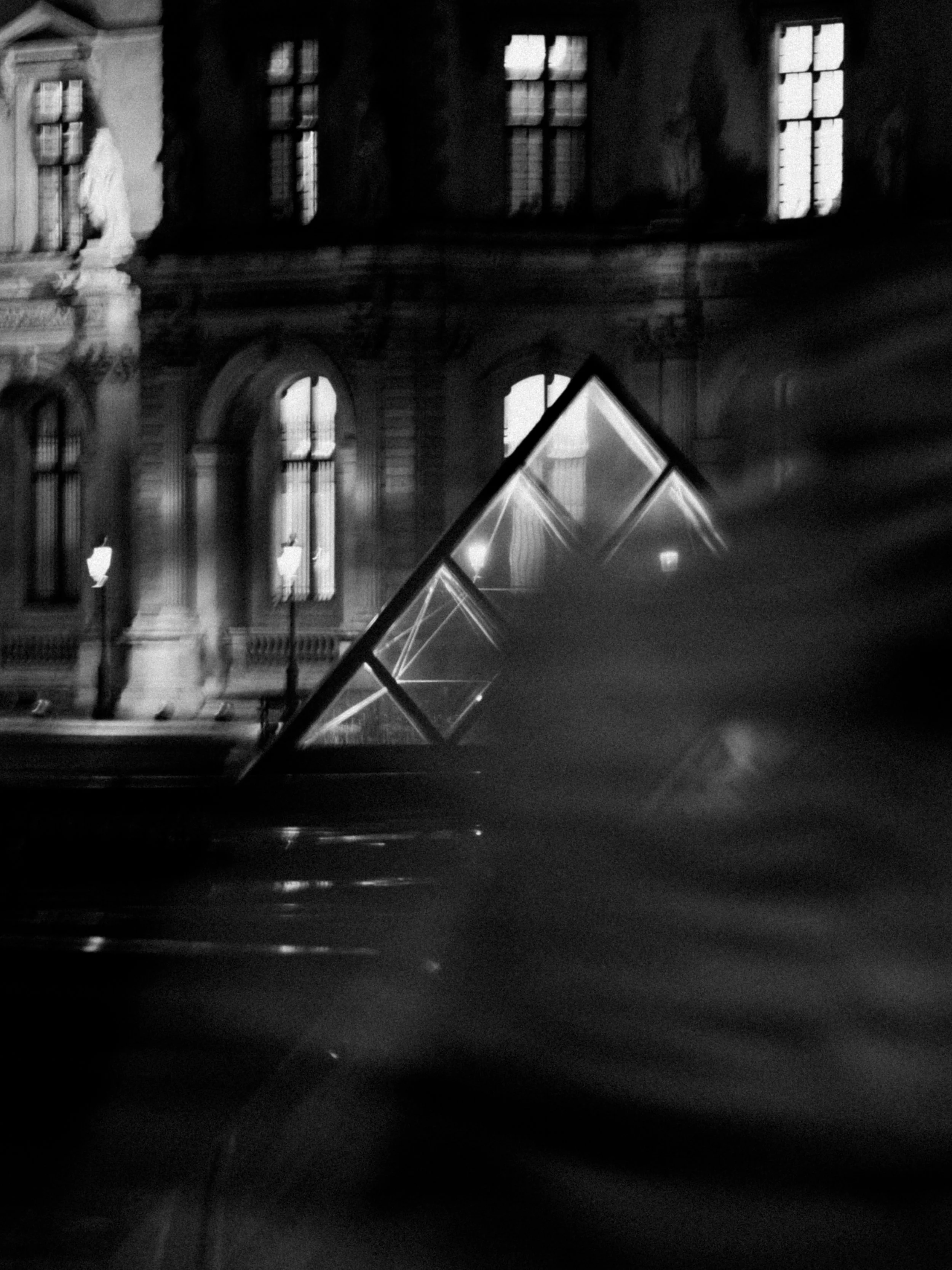 A black and white photo of a historic building with tall, lit windows and street lamps in the foreground. There is a reflective glass structure in the middle of the scene, possibly part of a modern installation or sculpture.