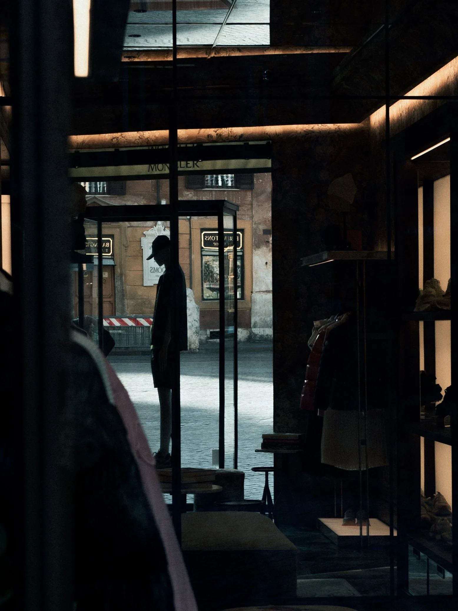 View through the glass door of a clothing store showing silhouettes of two people outside. The street scene includes buildings with signs and some greenery reflections on the glass.