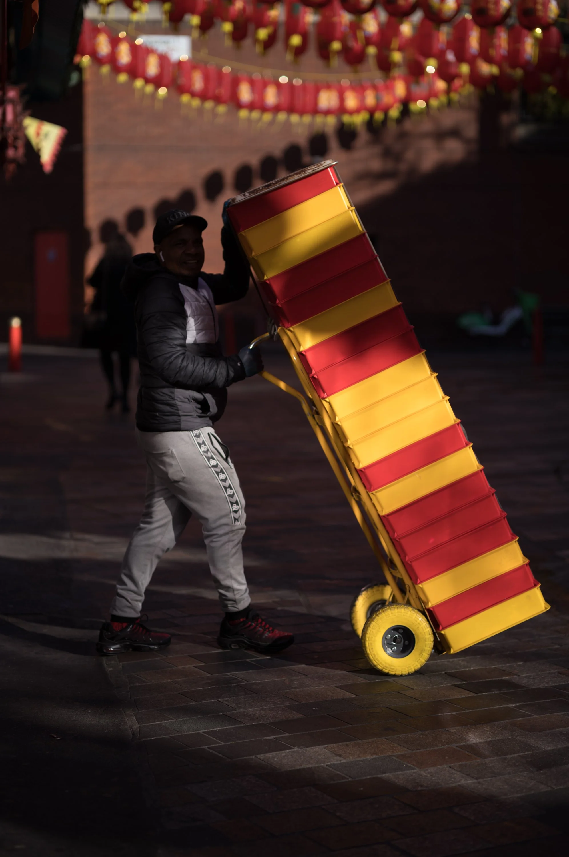 A person wearing a black jacket, gray pants with a pattern, and black shoes with red accents is pushing a yellow and red cart uphill on a dark, paved street.