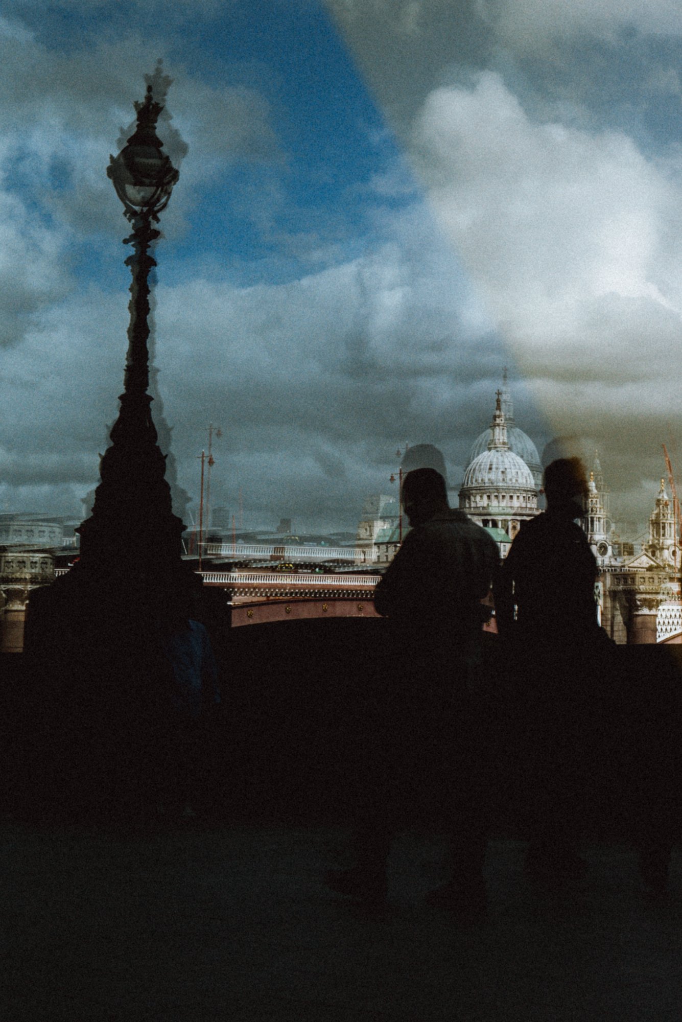 Silhouettes of two people standing on a bridge with the dome of St. Paul's Cathedral in London in the background, reflections and cloudy sky overhead.