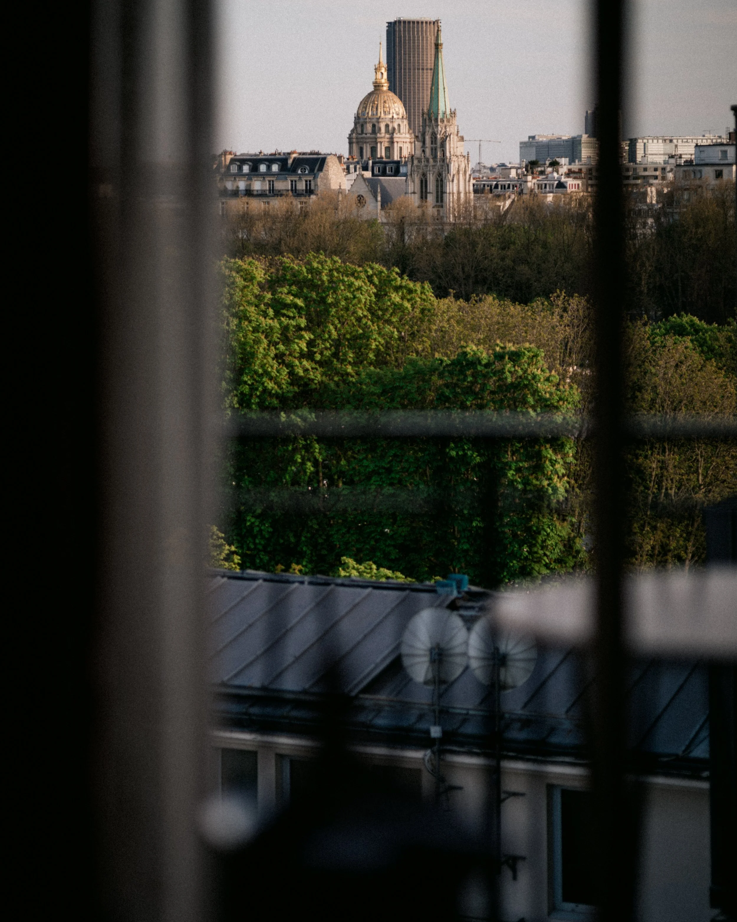 View of a city skyline through window blinds, including a historic building with a dome and tall spire, and modern high-rise buildings in the background.