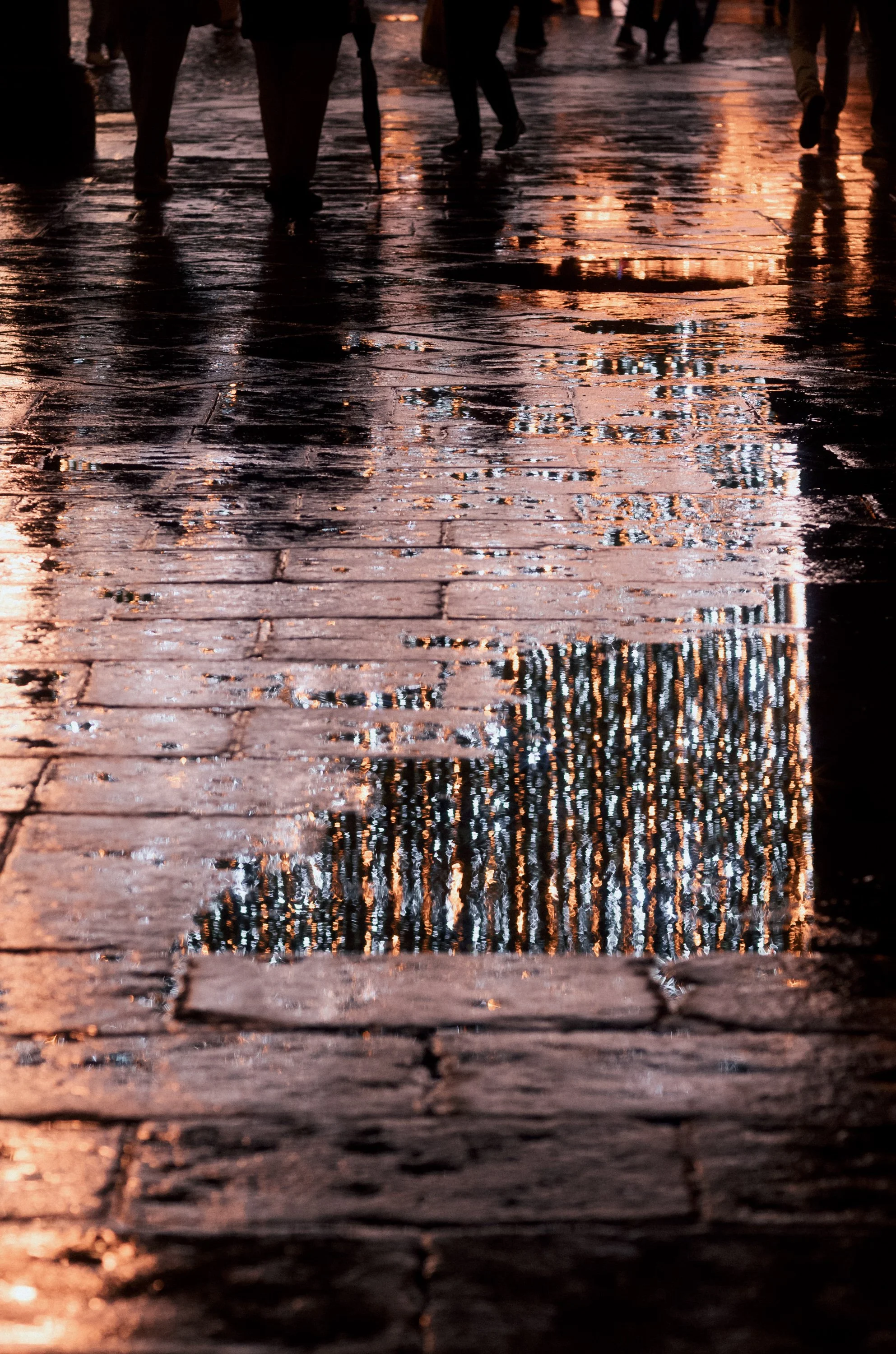 Reflection of a tall building with many windows on a wet brick sidewalk, with people walking in the background.