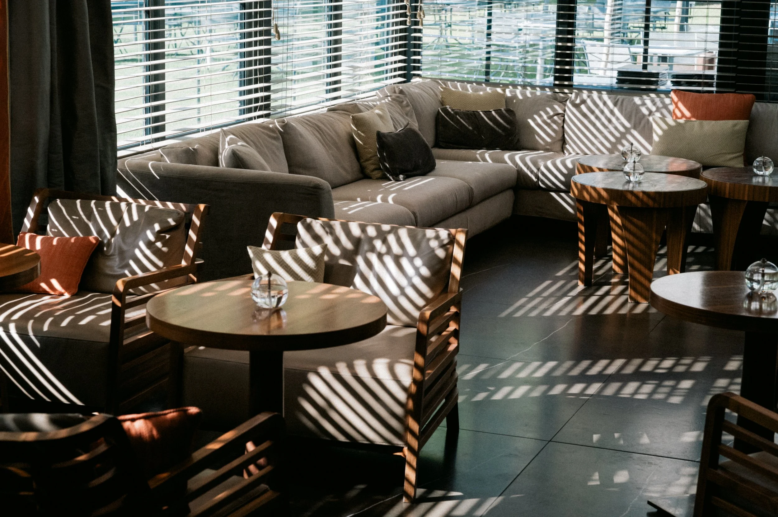 Interior of a modern living room or lounge area with a large sectional sofa, wooden tables, and chairs. Sunlight streams in through window blinds, casting striped shadows across the furniture.