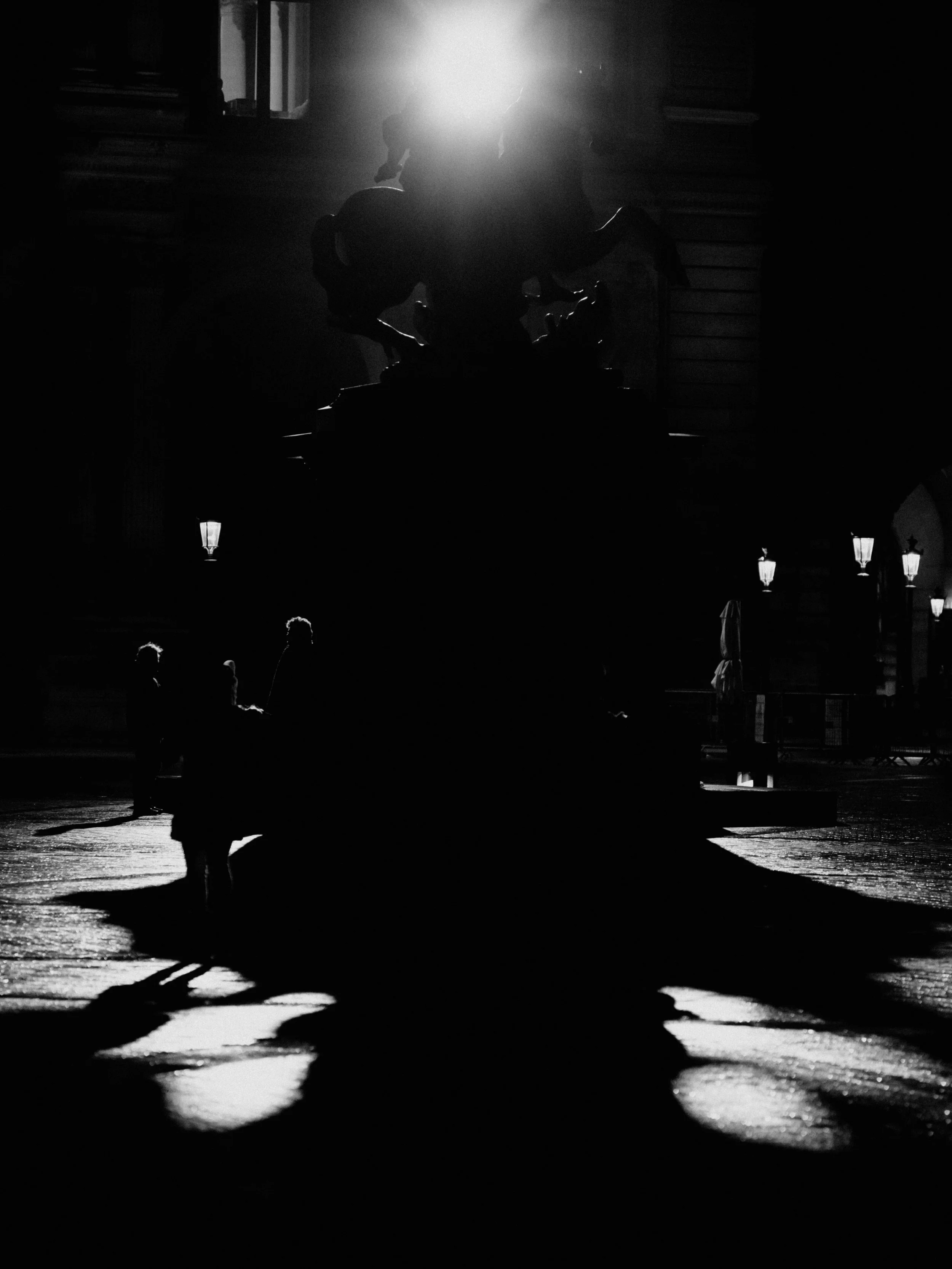 Silhouette of a statue with a horse and rider in front of a light source, surrounded by smaller figures and street lamps at night.