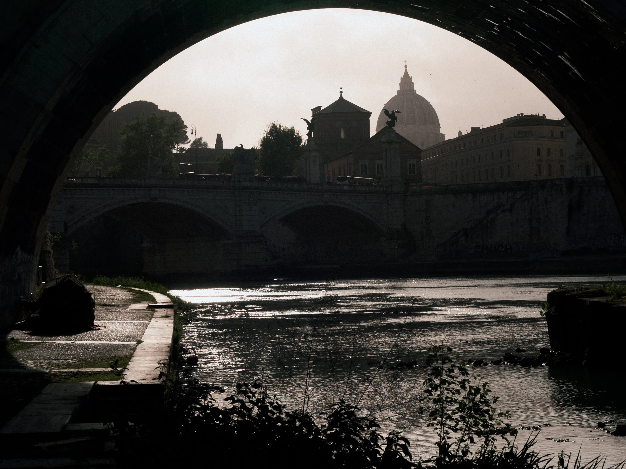 Cityscape viewed through an arch bridge, with a river in the foreground reflecting sunlight, buildings, trees, and a prominent domed structure in the background.