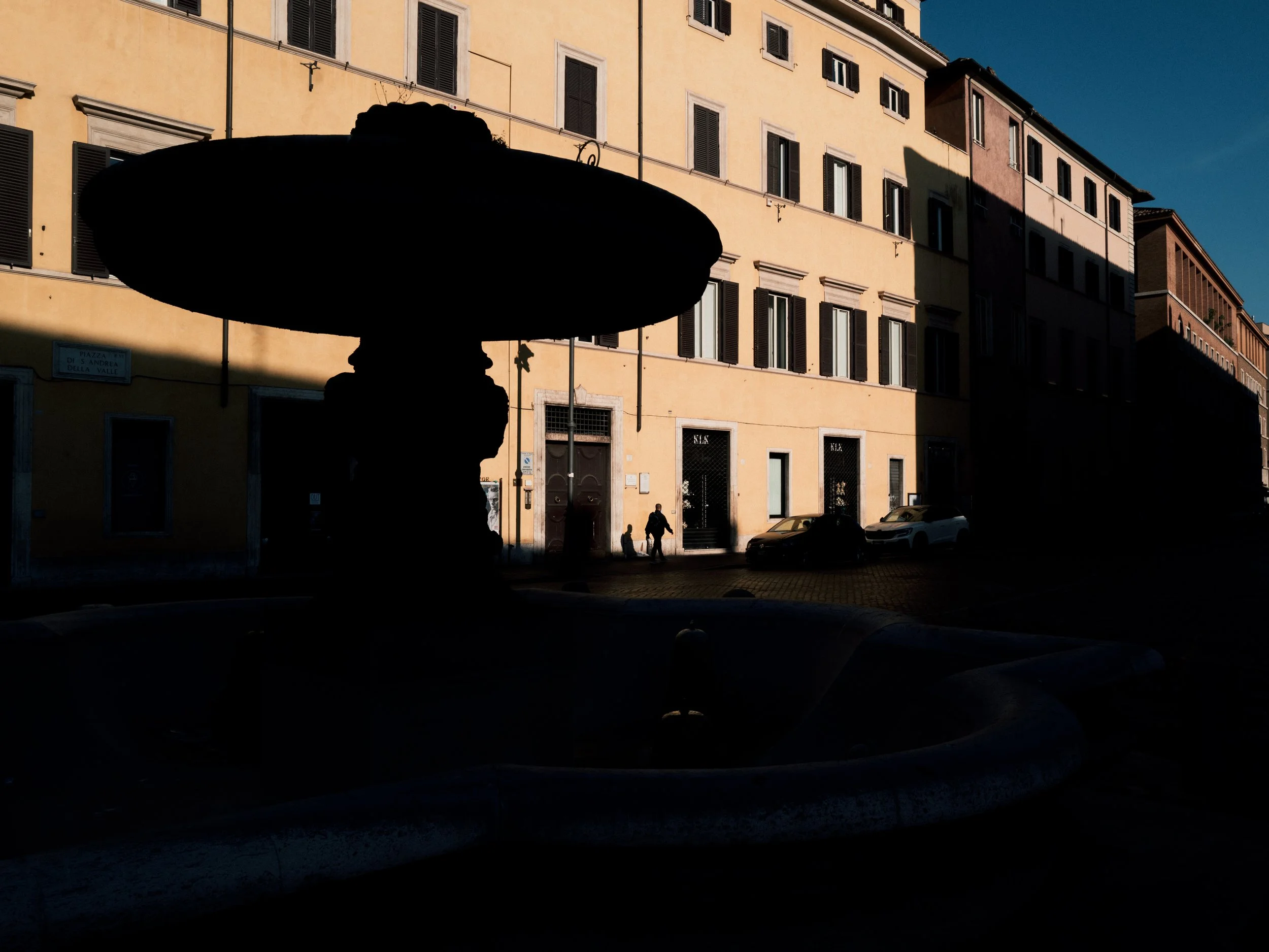 Silhouette of a person wearing a hat in front of a sunlit building with multiple windows and closed shutters, with a couple walking and parked cars visible on the street.
