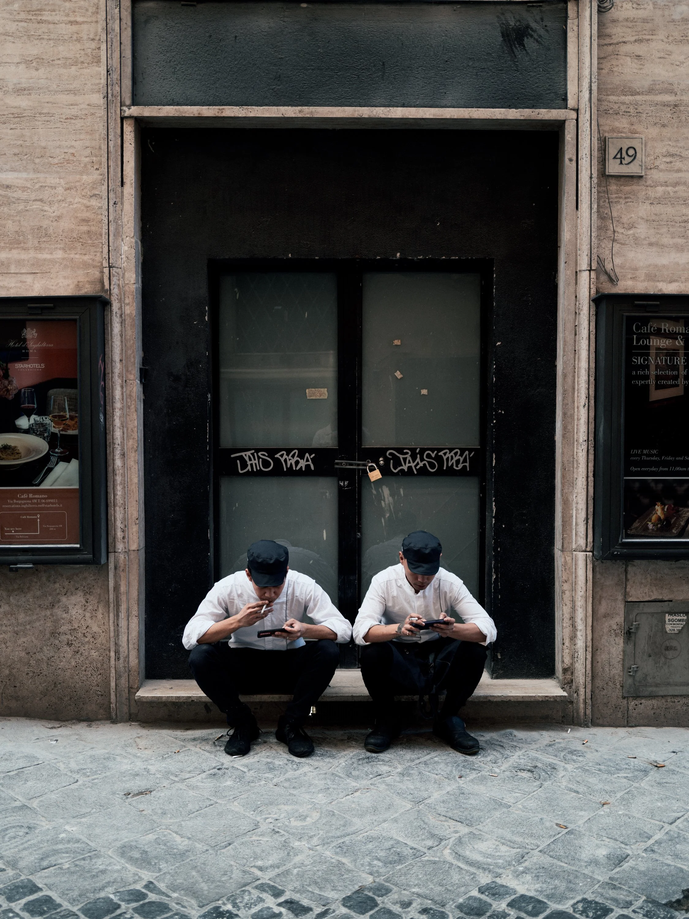 Two men wearing white shirts, black pants, and black caps are squatting on a stone sidewalk in front of a closed black door with glass panels, out of which graffiti is visible. They are looking at their phones.