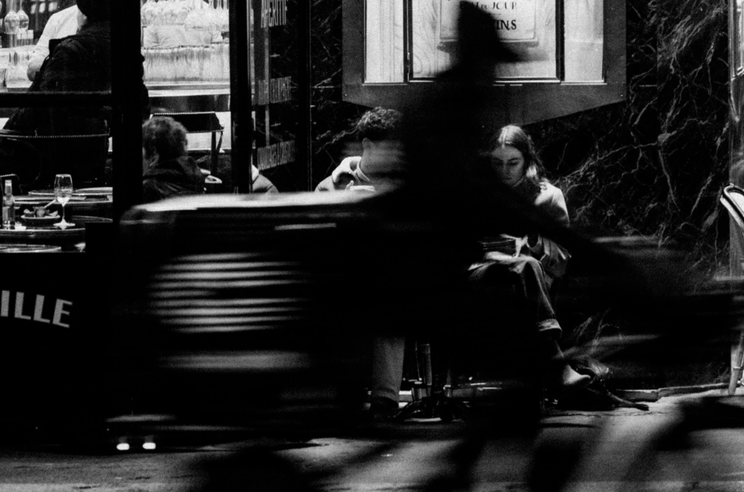 A black and white photo depicting a nighttime scene at an outdoor cafe or restaurant with several people sitting at tables, some engaged with their phones, and blurred silhouettes of pedestrians walking by.