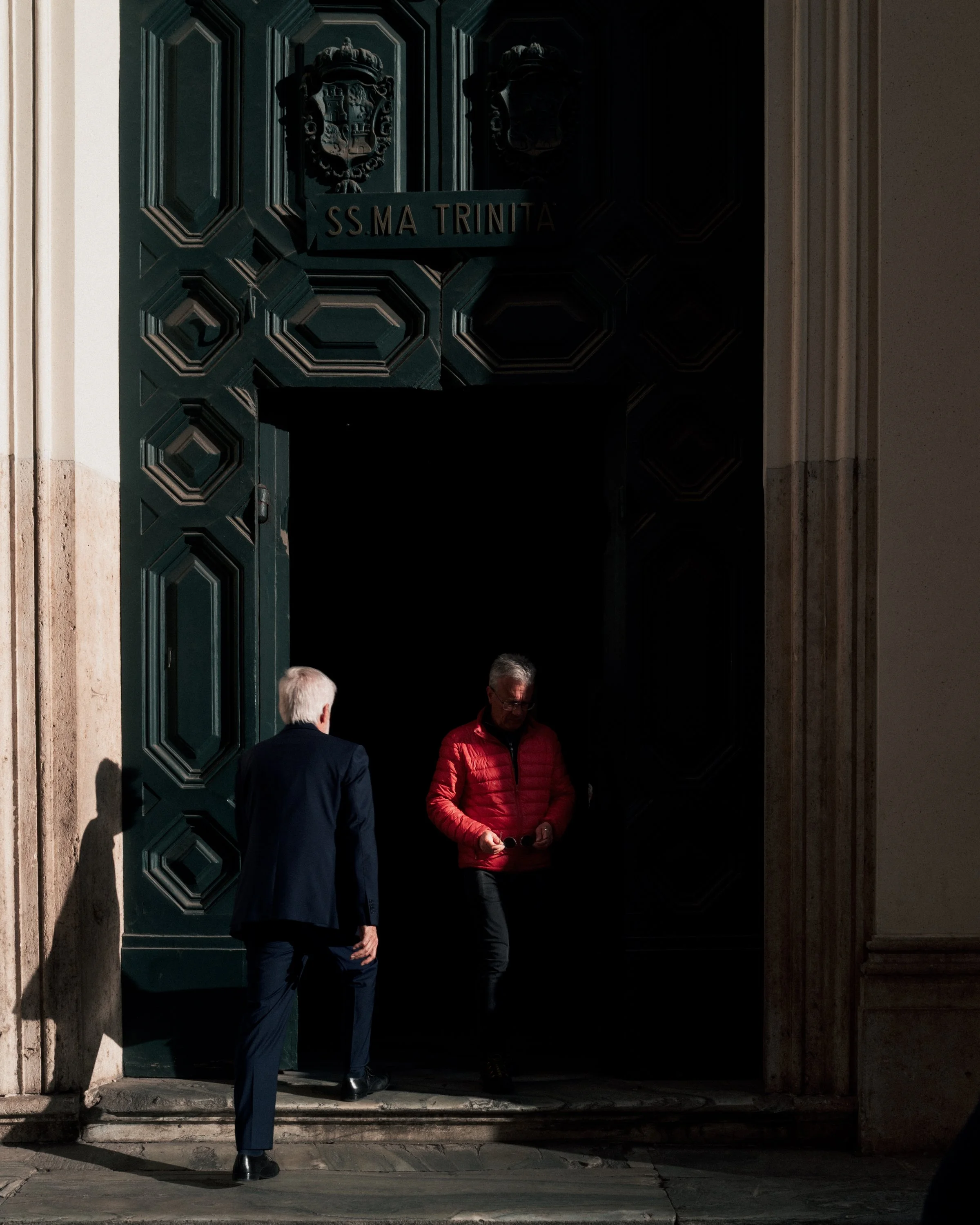 People standing at the entrance of a dark building with a large green door with shields and the words S.S. MA Trinità on a banner above the door.