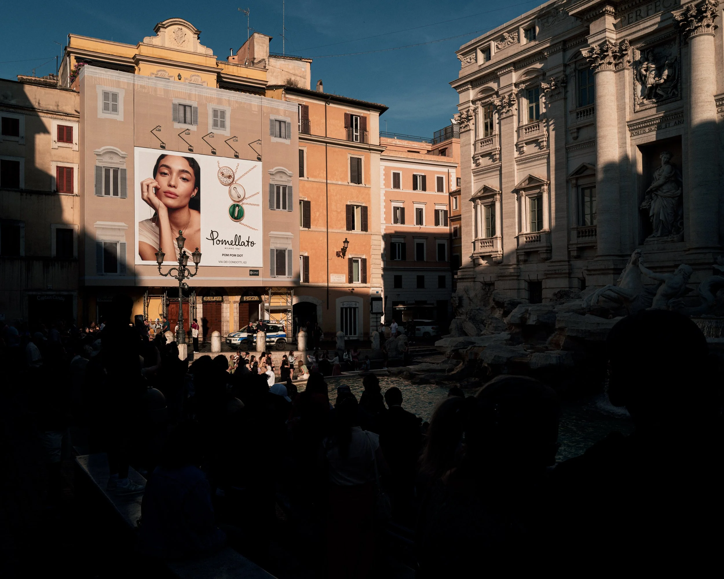 A city square with historic buildings, a billboard showing a woman's face and jewelry advertisement, and a fountain with people sitting around it. The buildings are in warm pastel colors, and the scene is lit by late afternoon sunlight.