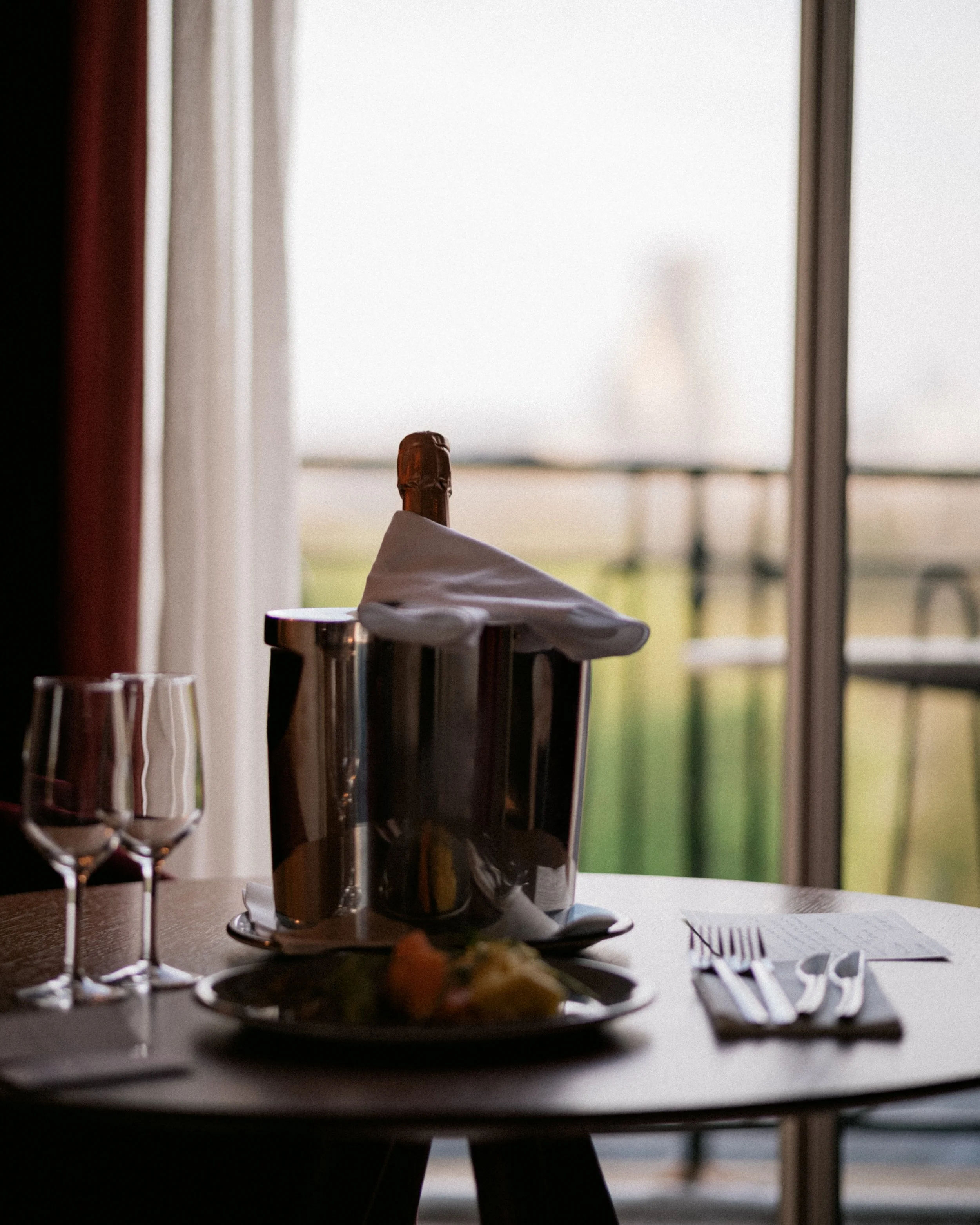 A table set for dining with a bottle of liquor, glasses, and utensils. A window with curtains in the background shows an outdoor scene.