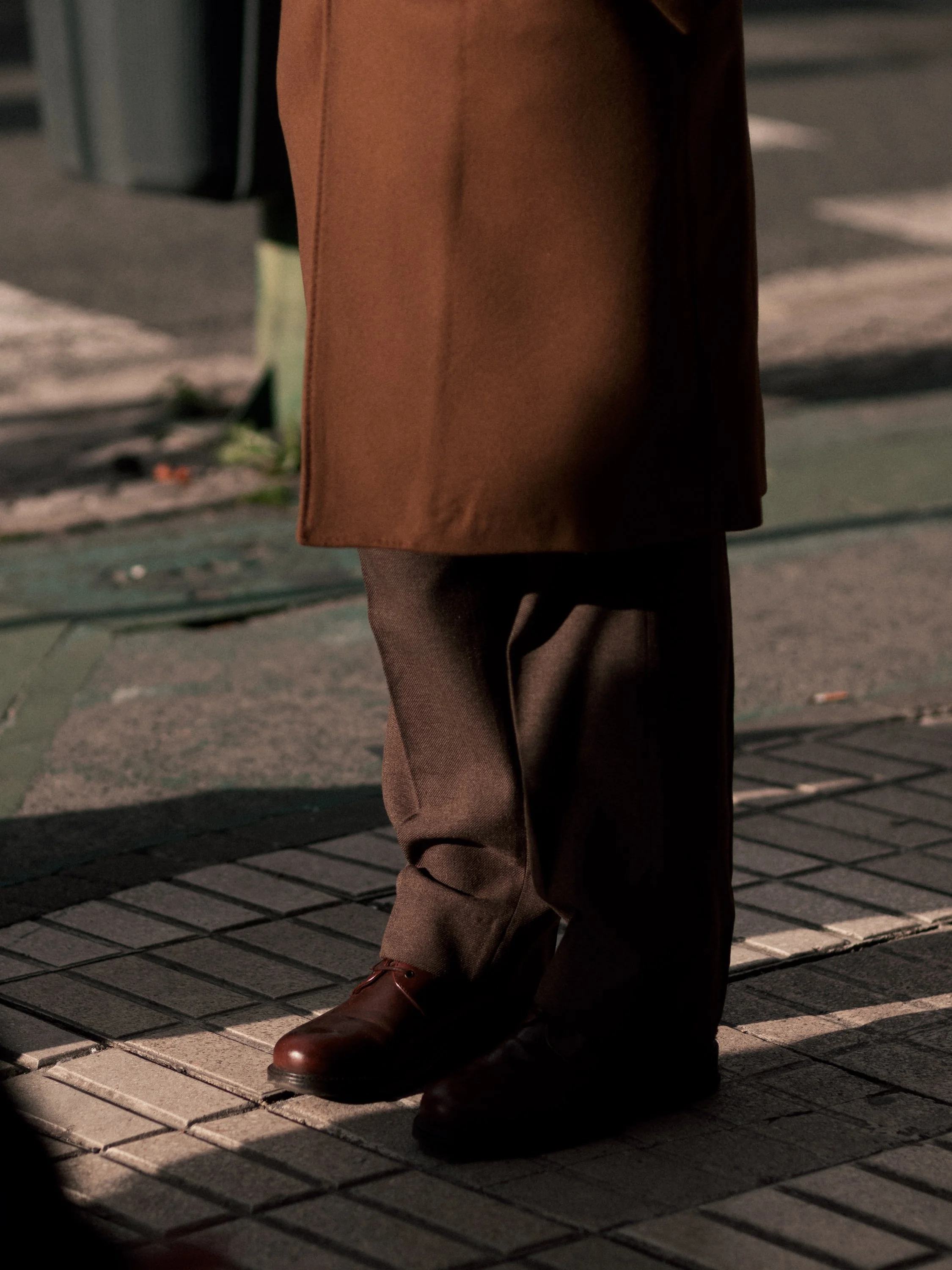 Close-up of a person's legs and feet standing on a sidewalk at night, wearing brown dress shoes, dark trousers, and a brown coat.