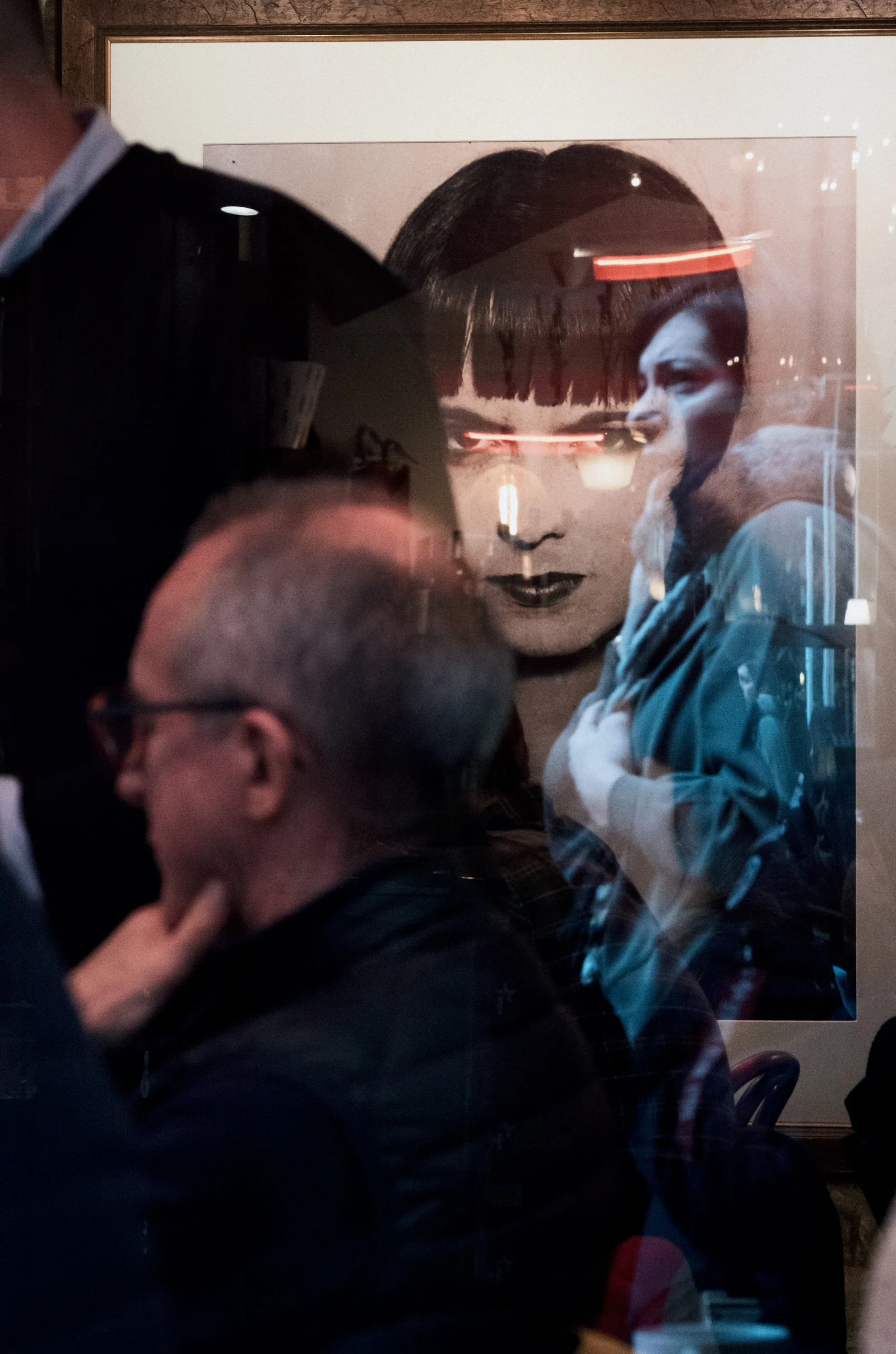 Reflected image of a mall portrait of a woman with dark hair and bangs, and a man with glasses, in a busy indoor setting with people and lights.