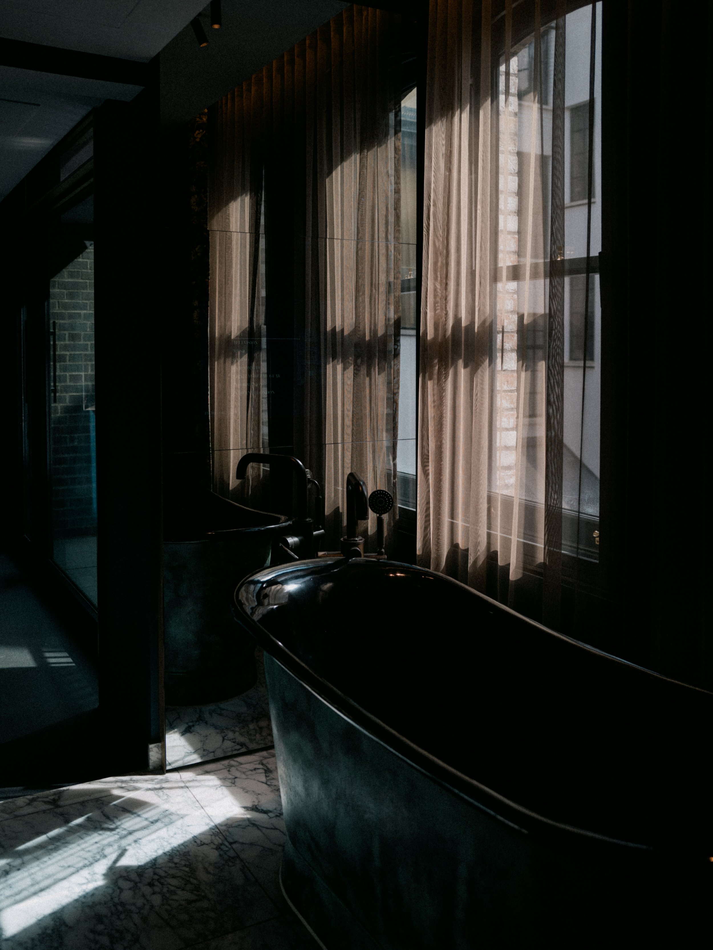 Dark interior space with large window curtains filtering sunlight onto marble floor and dark basin in foreground.