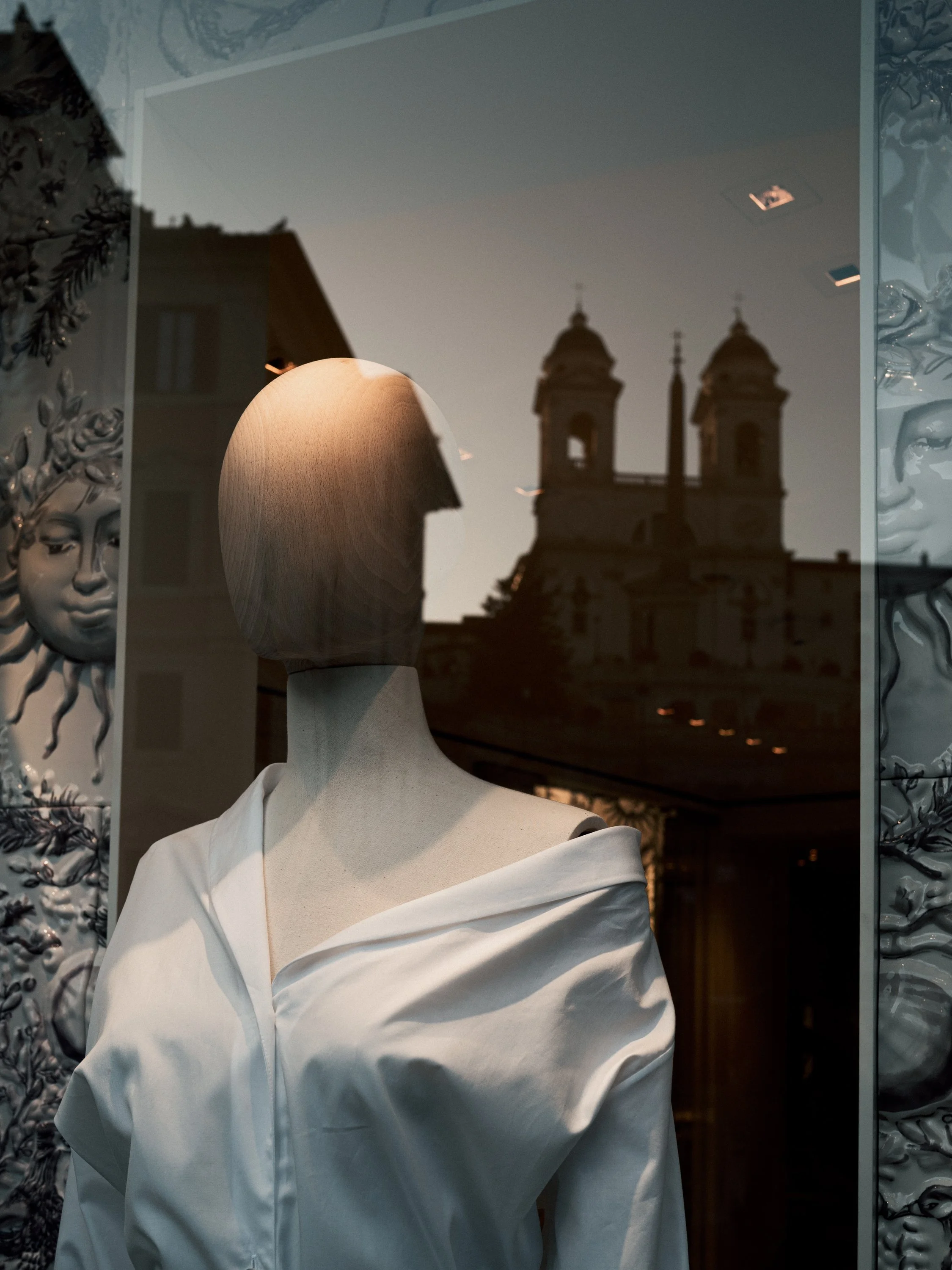A mannequin wearing a white off-shoulder blouse standing in front of a shop window. The window reflects a building with two towers and a clear sky.