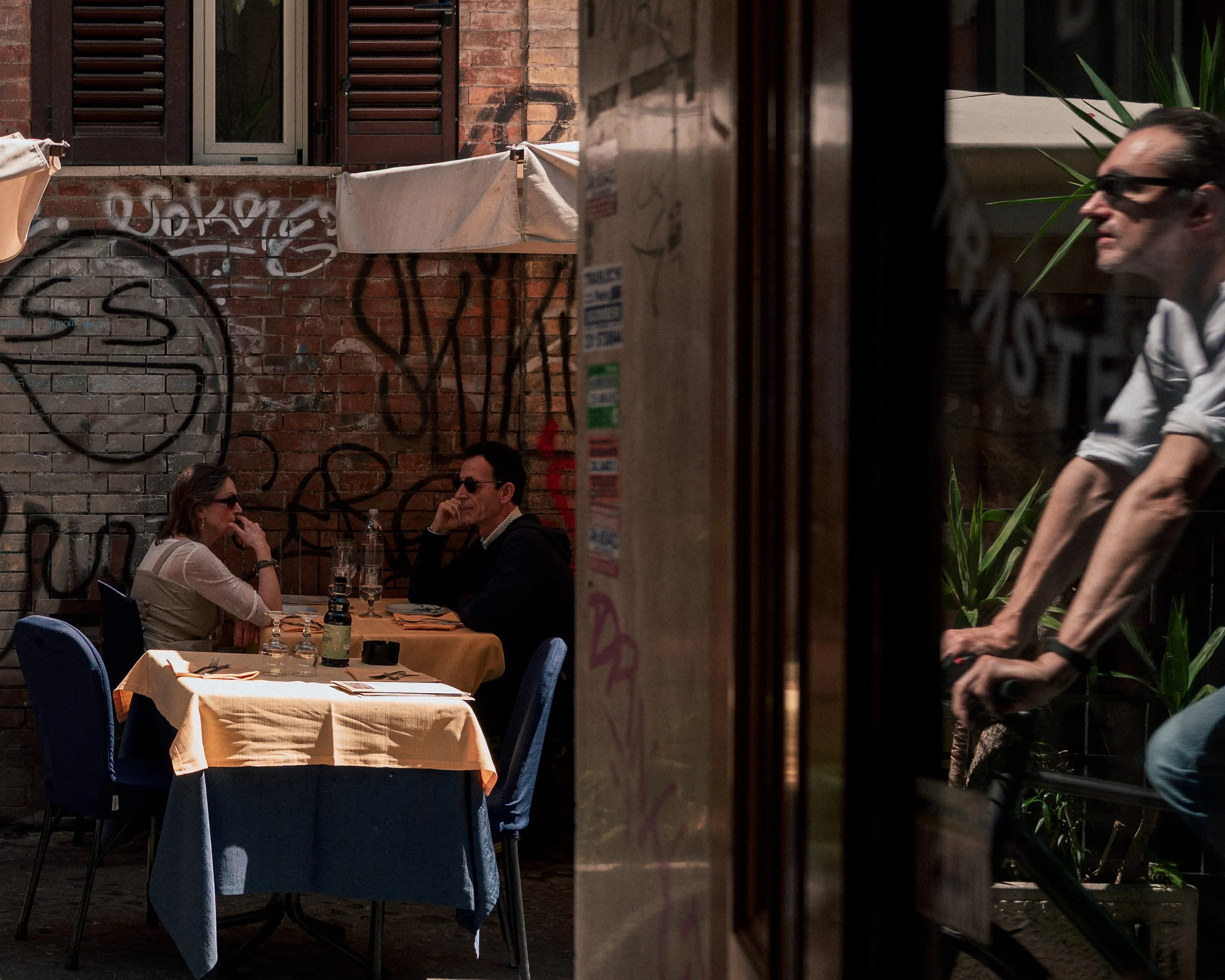 People dining outdoors at a restaurant, two people are sitting at a table covered with a yellow tablecloth, one woman and one man are visible, and a server is walking past.