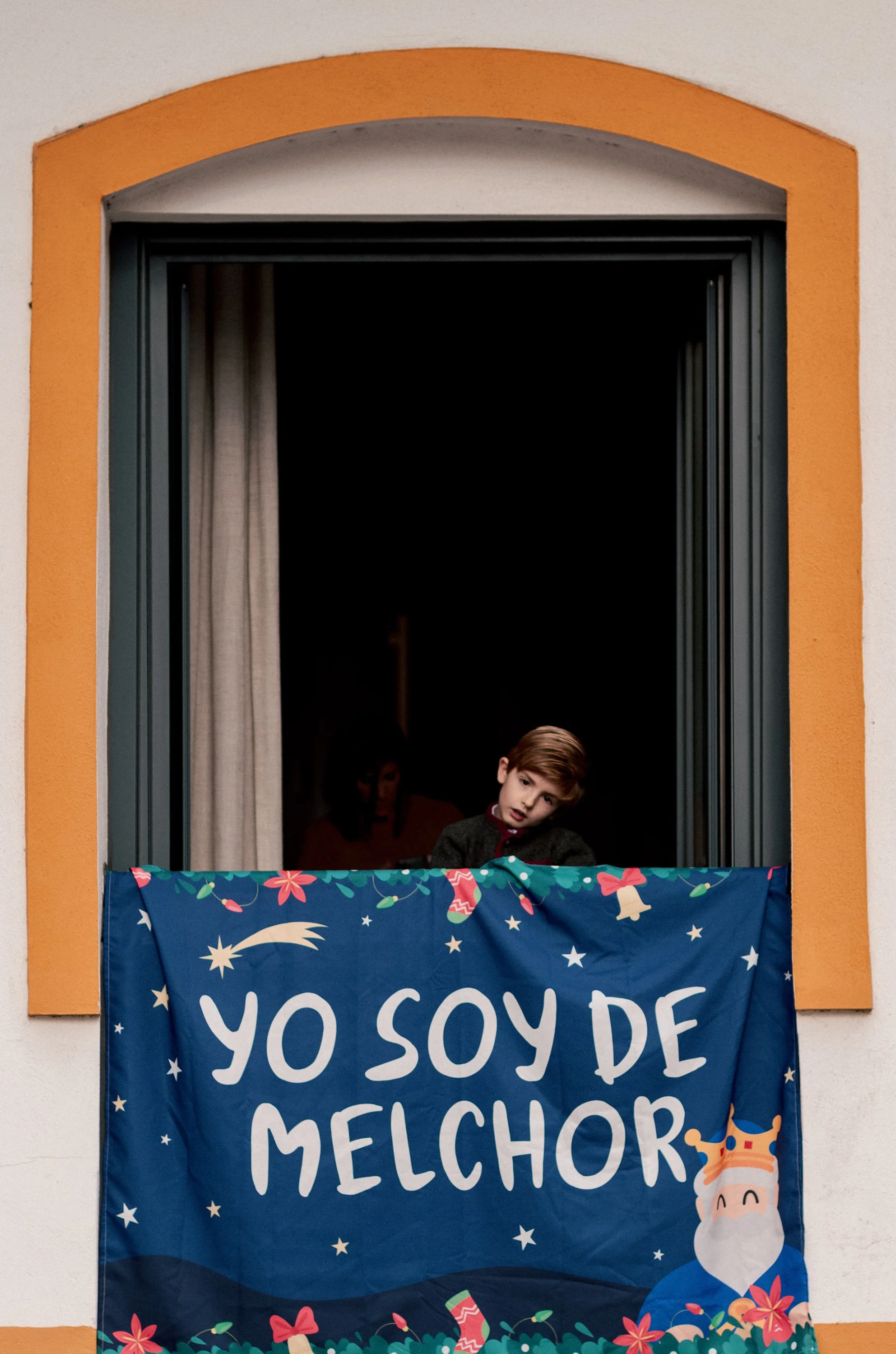 A young boy looking out of a window with a Christmas-themed banner that reads 'Yo soy de Melchor,' with festive decorations and Santa Claus illustration.