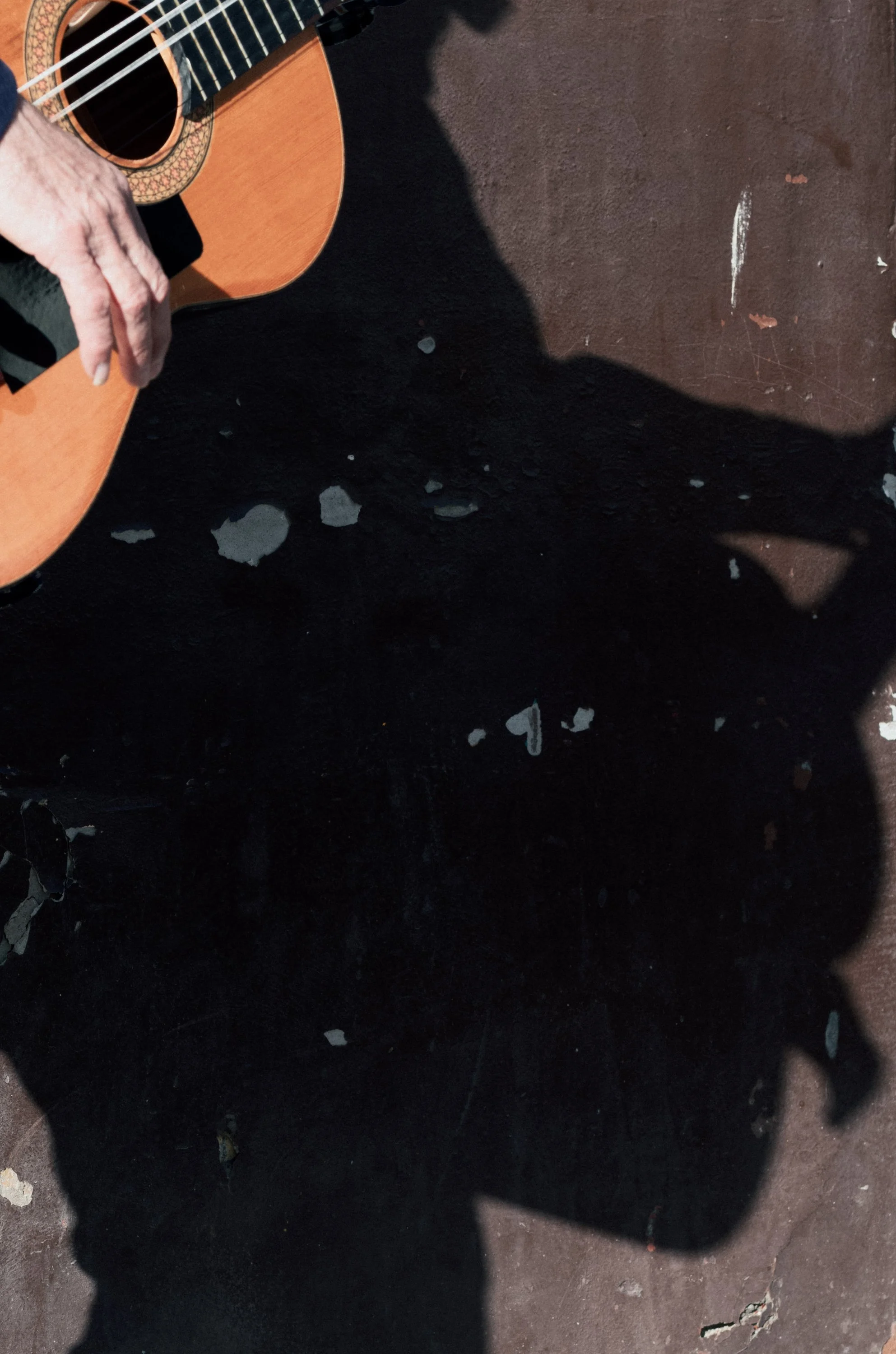 Close-up of a person’s hand playing an acoustic guitar, with a large shadow cast on the ground.