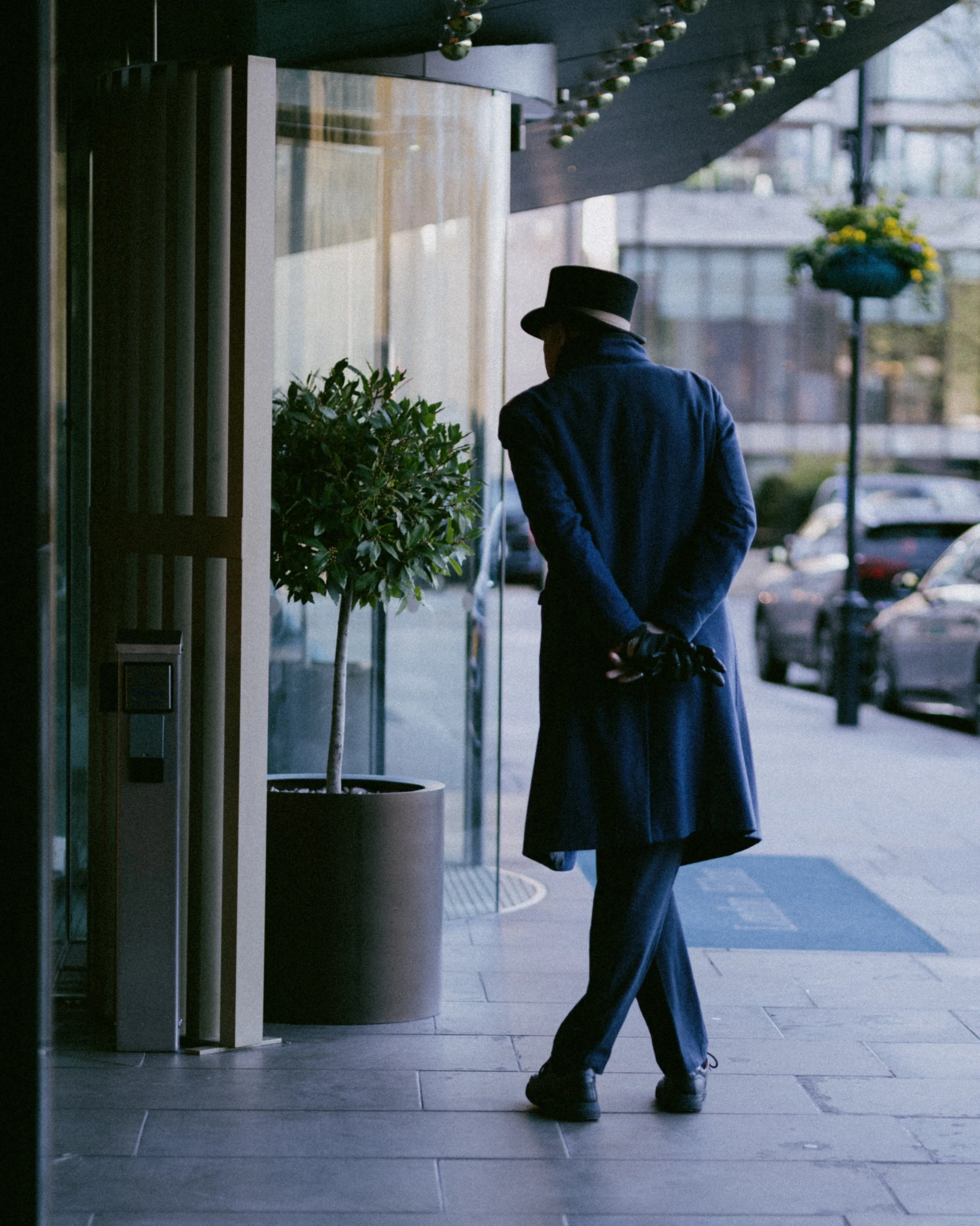 Person in a dark coat and hat waiting outside a building, standing near a potted plant with cars parked on the street in the background.