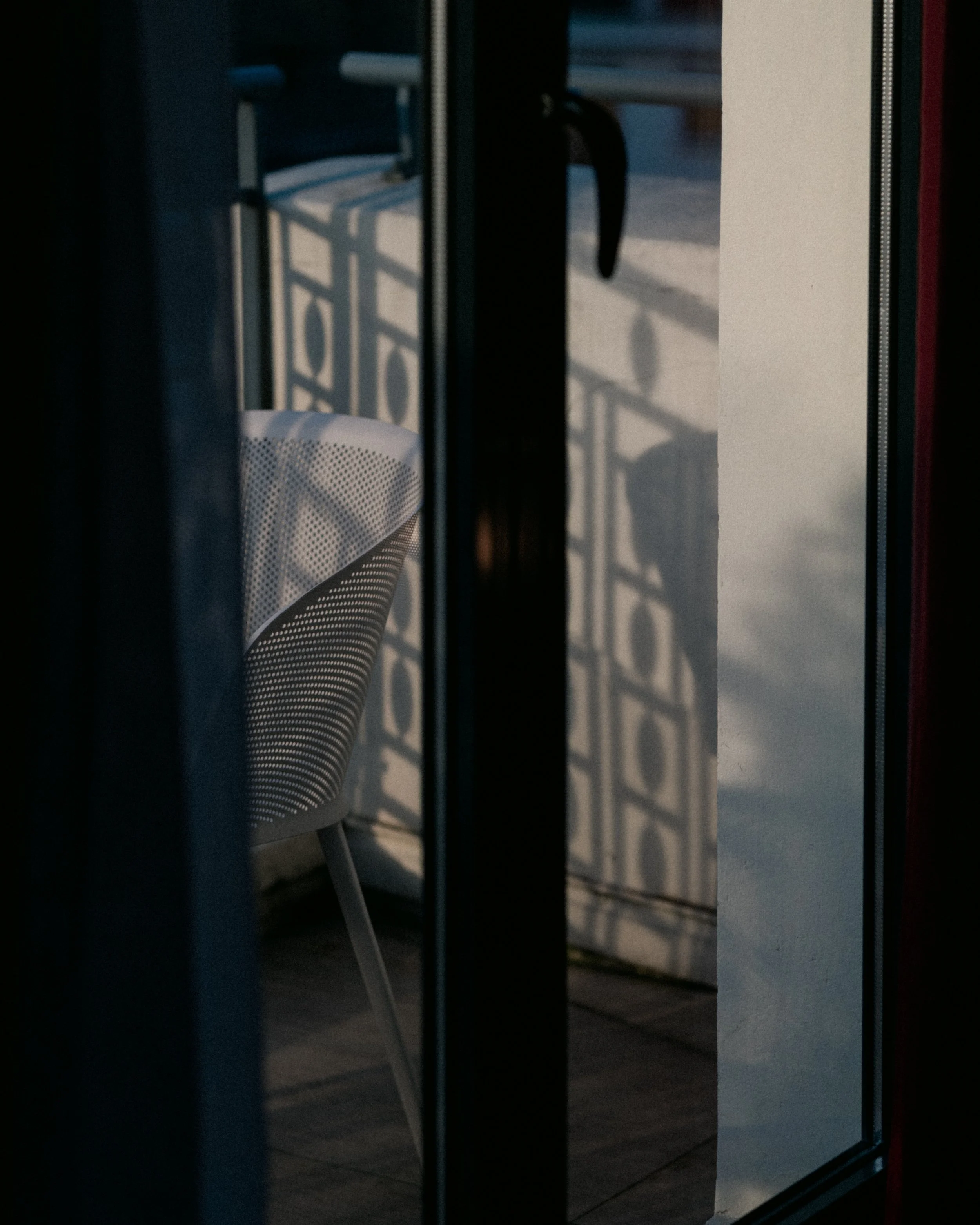 A partially open glass door shows a white plastic chair outside, with sunlight casting a shadow of metal railing on the ground and the chair.